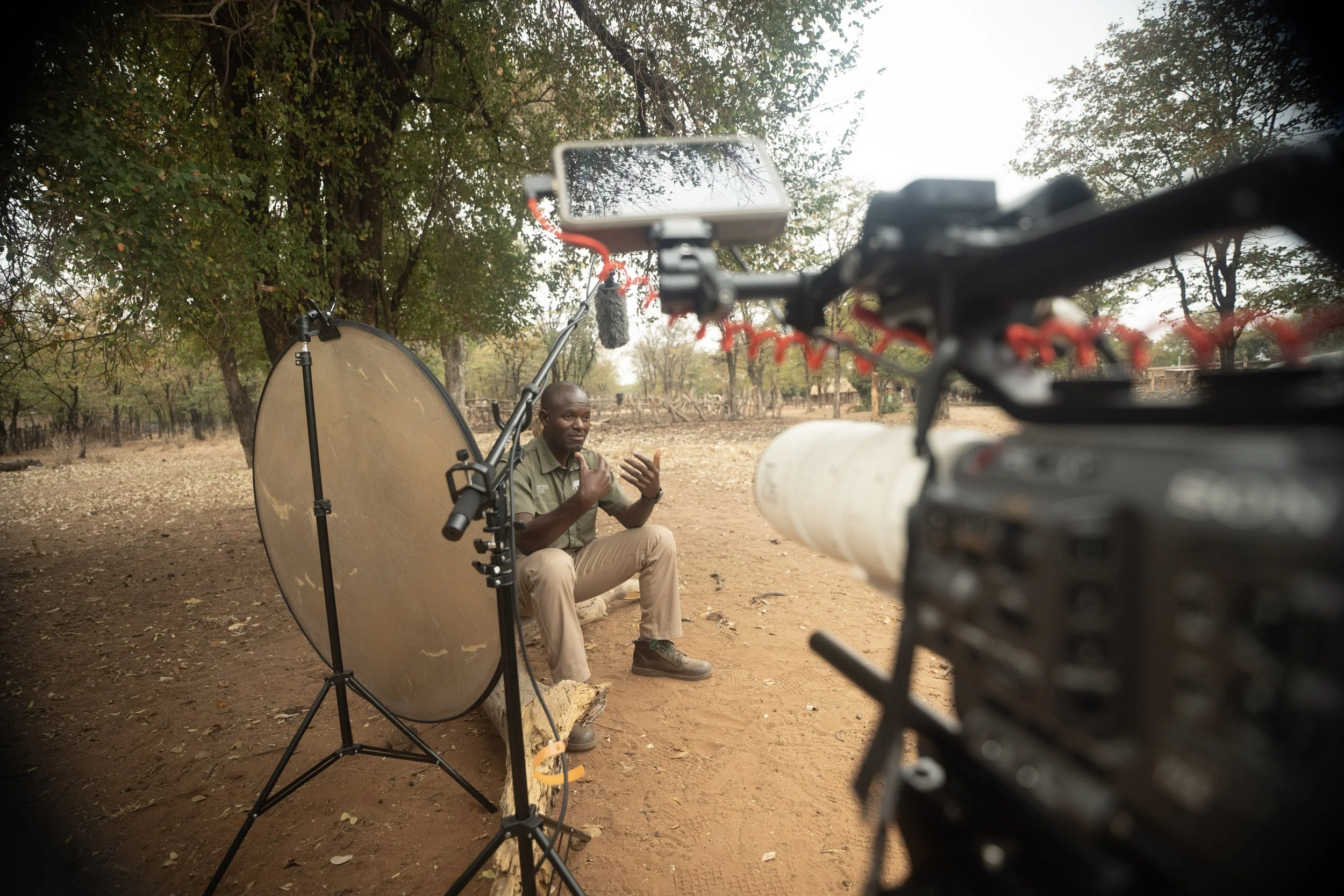 Man sitting outdoors on dirt ground, speaking in front of camera equipment with a ring light, backdrop of trees and open land.