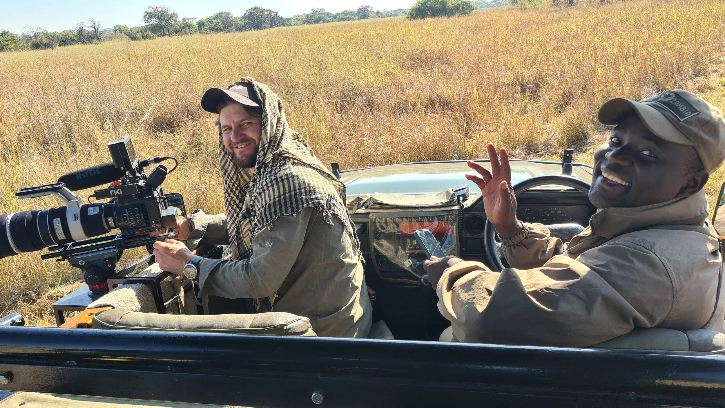 Two men are on a safari game drive in an open vehicle, surrounded by tall grass. One man, holding a camera with a large telephoto lens, is smiling at the camera. The other man, sitting beside him, is wearing a beige hat and jacket, smiling and making