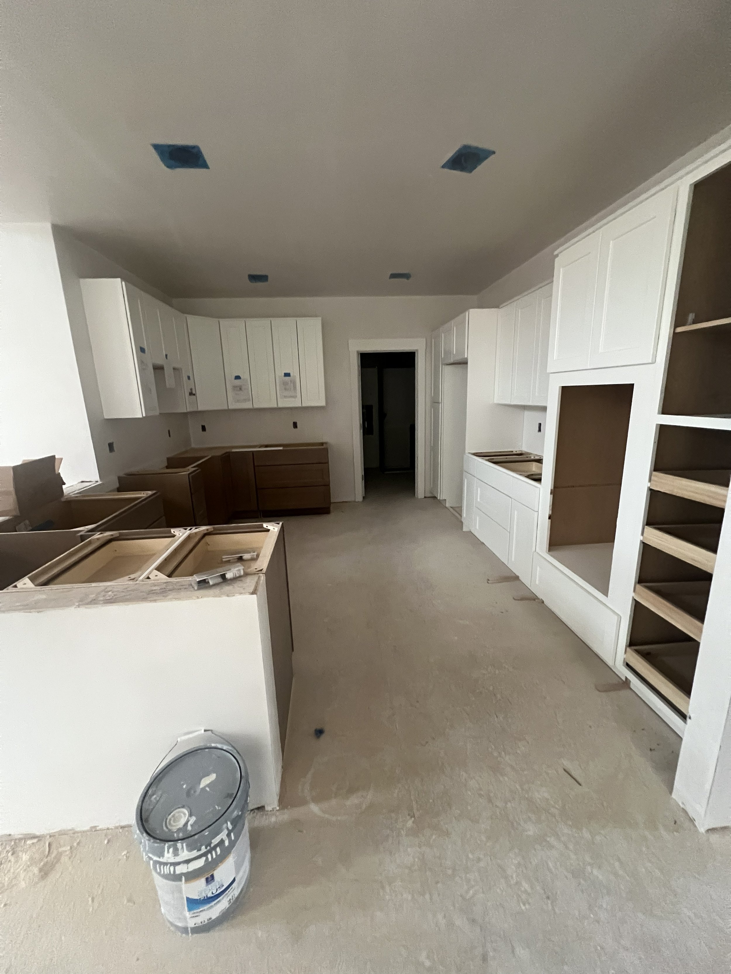 Kitchen under construction with white and brown cabinets, open shelving, and a bucket on the unfinished floor.