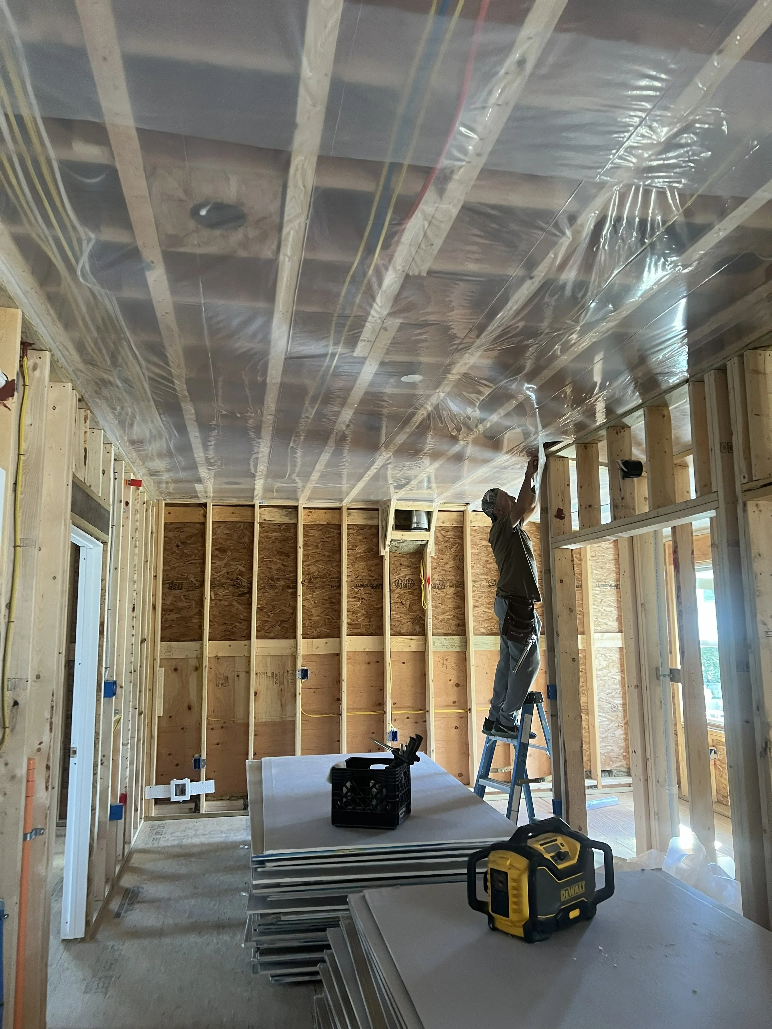 A construction worker standing on a ladder installing a plastic vapor barrier on the ceiling of a framed room under construction.