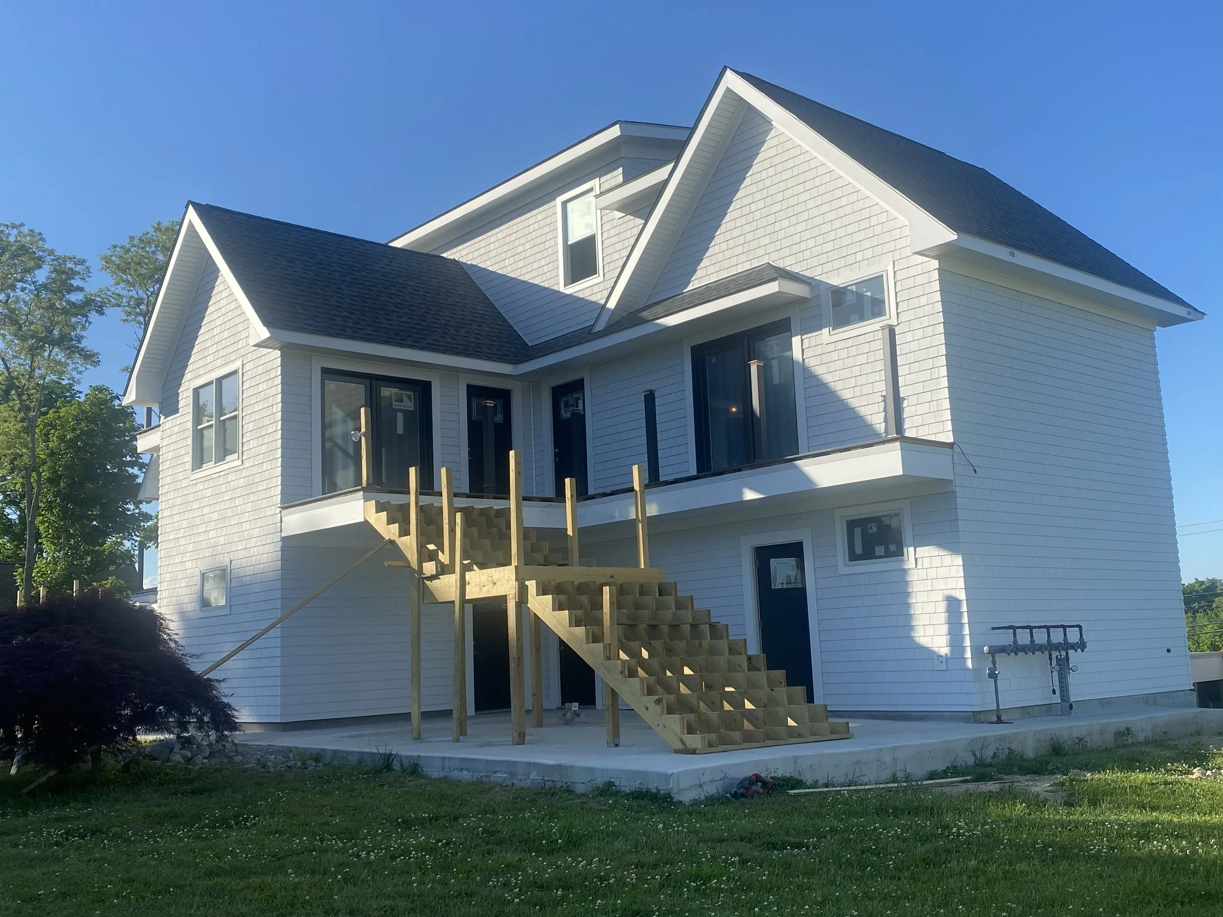 A two-story house under construction with wooden stairs leading to a balcony. The house has white siding, multiple windows, and a dark gray roof. The front yard has green grass and a small tree.
