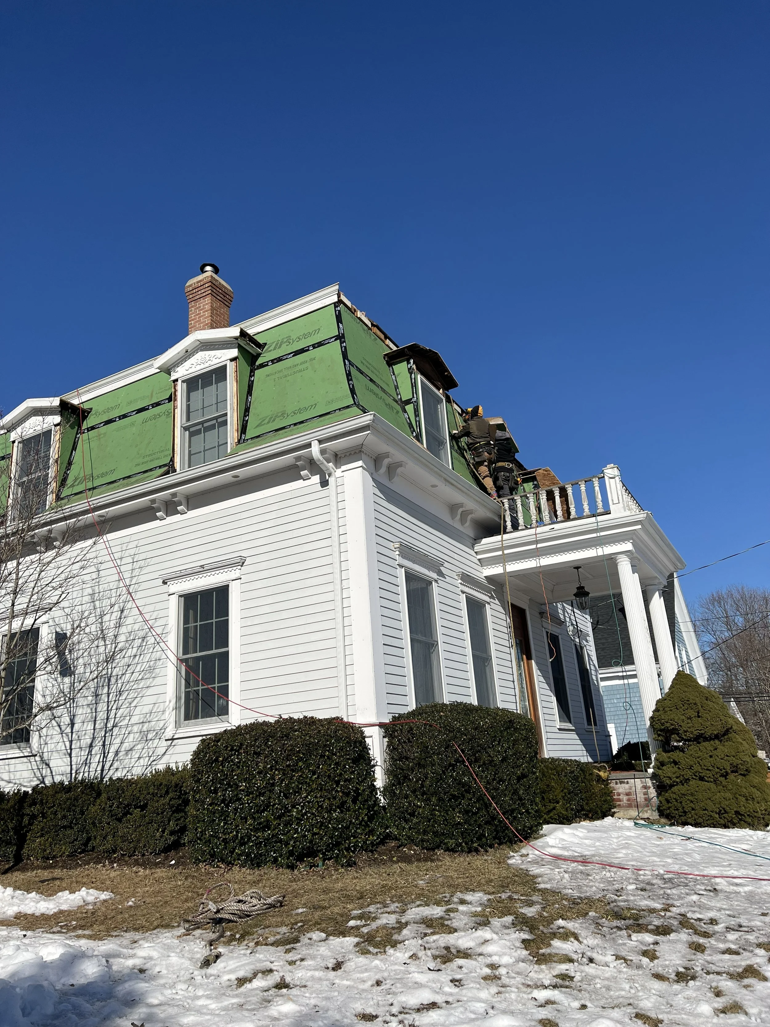 Workers renovating or repairing the roof of a large white house with a porch, set against a clear blue sky, with some snow on the ground.