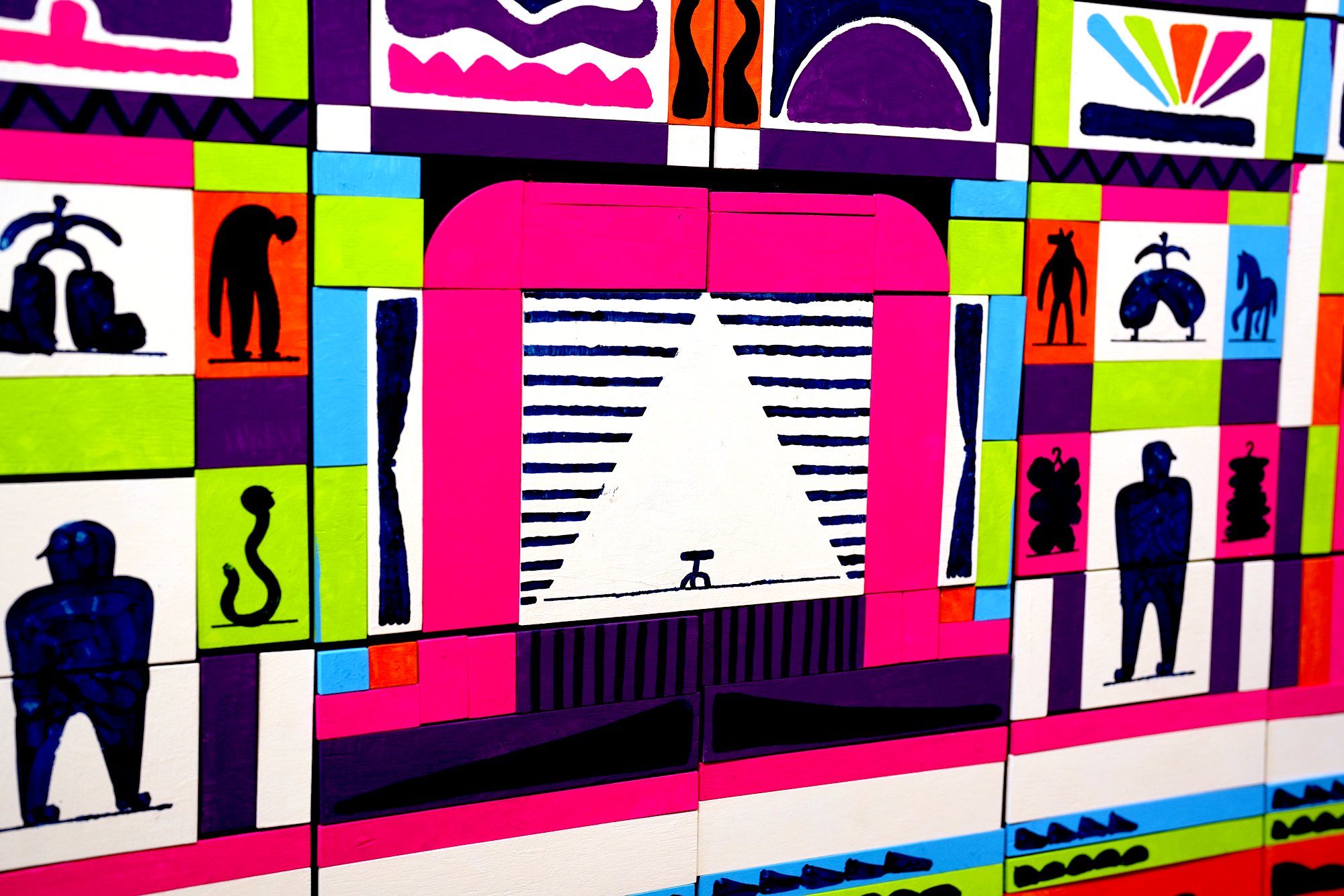 Colorful abstract art made of small blocks with various patterns and silhouettes, including a door-like opening, snake, animal, and human figures.