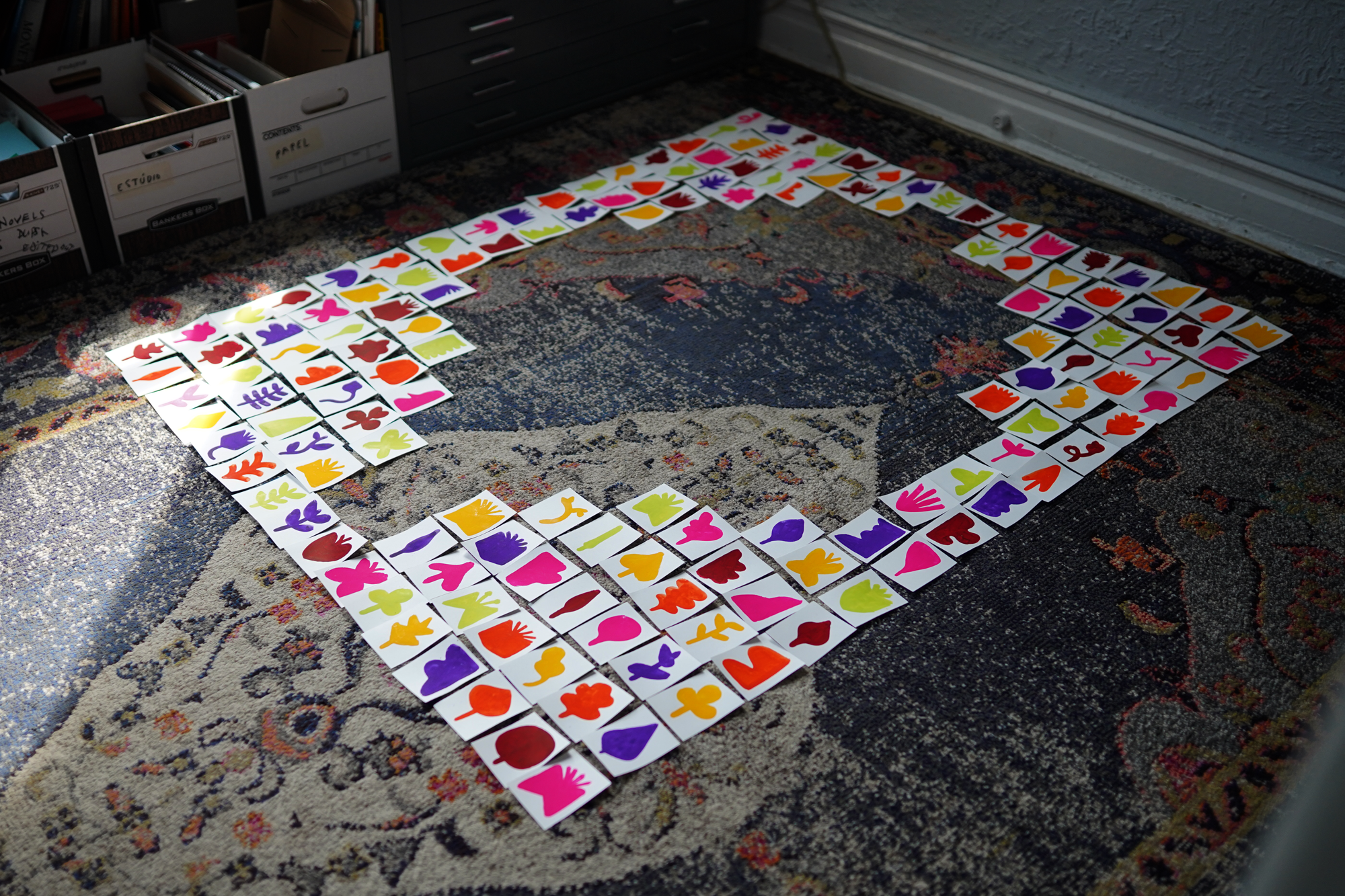 Colorful paper cutouts of various shapes and silhouettes arranged on a carpet in a square pattern.