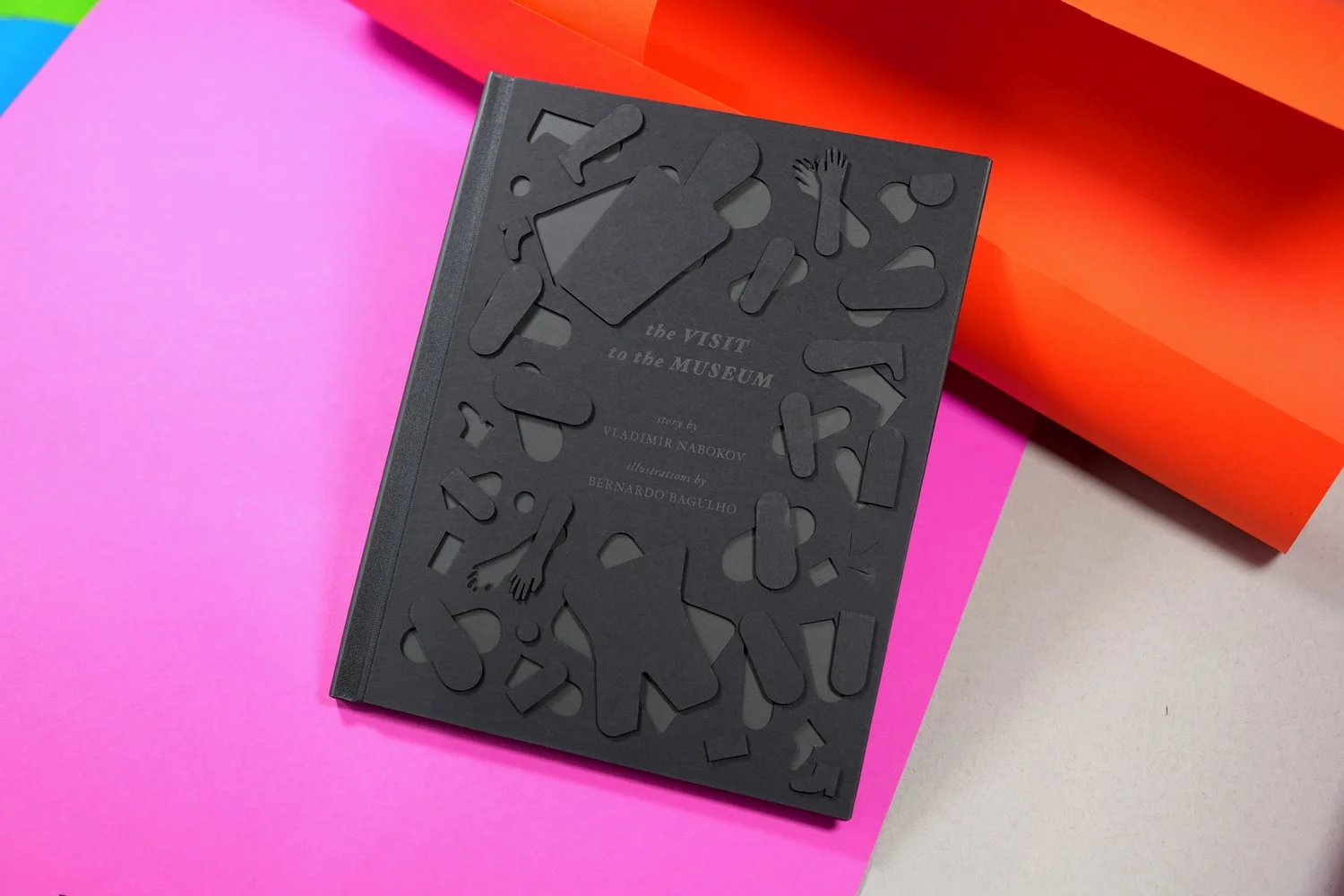 Gray book titled 'the VISIT to the MUSEUM' with embossed silhouettes of museum-related objects, placed on pink, gray, and orange backgrounds.