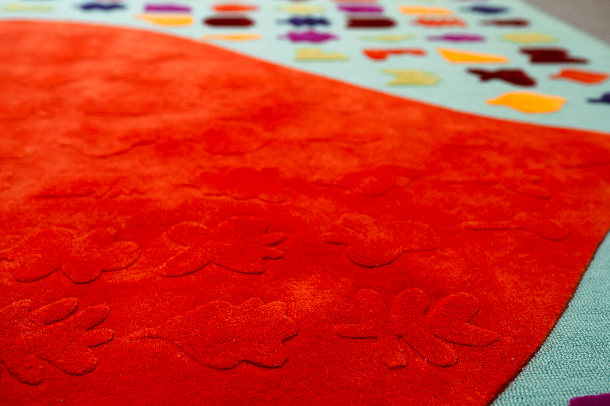 Close-up of a red textured quilted fabric with scalloped stitching, partially covering a colorful fabric with small, multicolored shapes or patterns.
