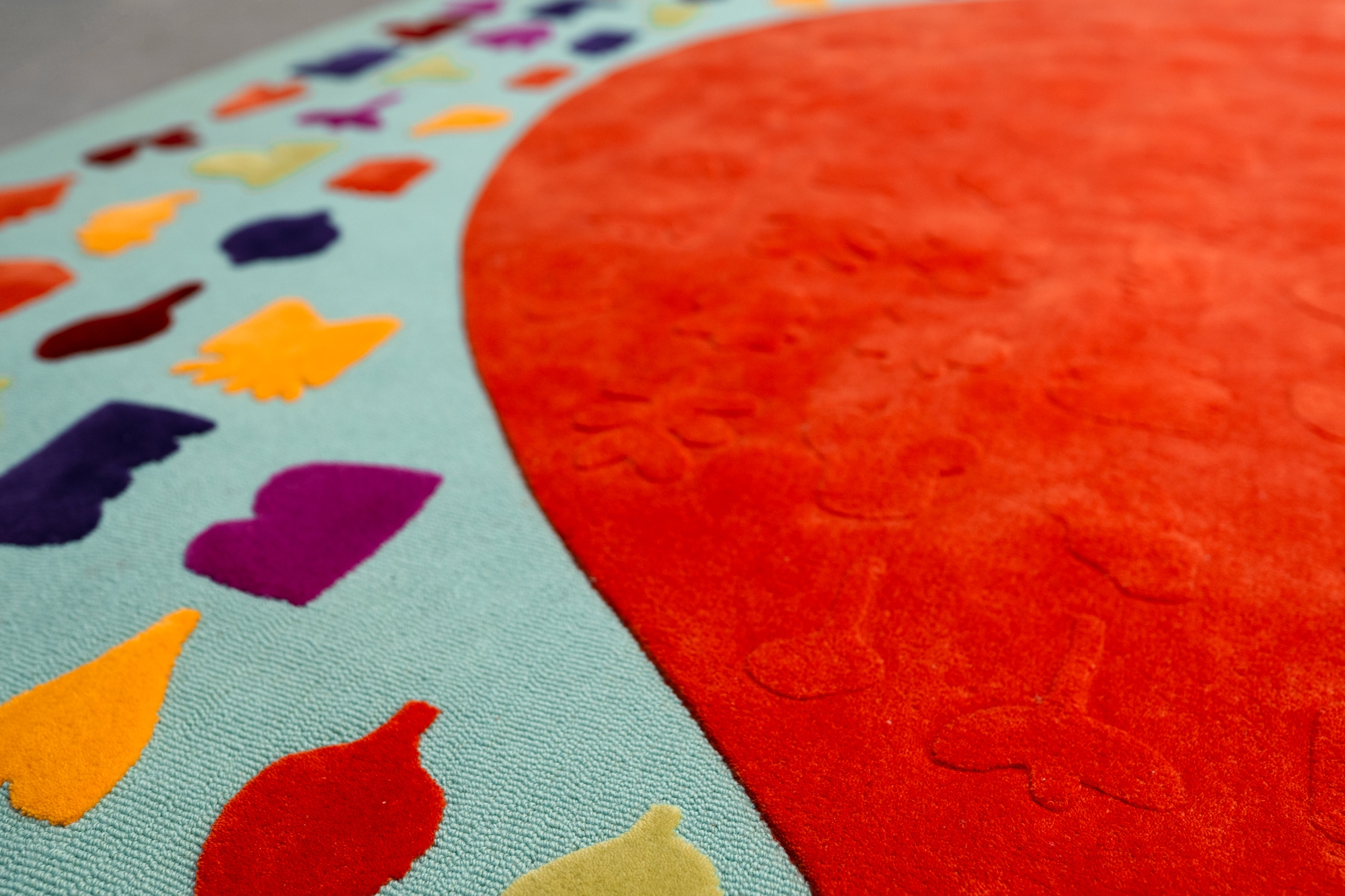 Close-up of a colorful rug featuring a large red circle and a border with various small, colorful shapes on a light blue background.
