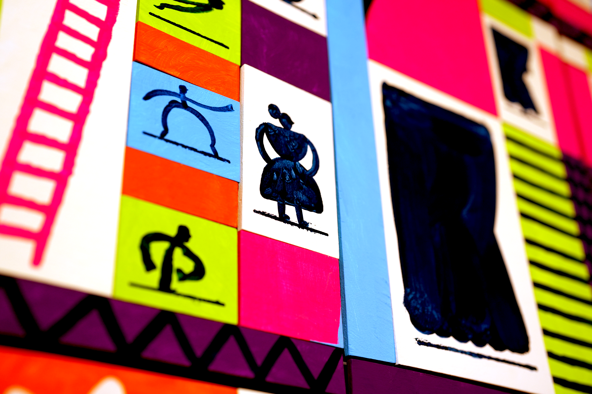 Colorful wooden blocks painted with various illustrations including a gymnast, a girl with a skirt, a person with a backpack, and a person in a running pose, arranged in a geometric pattern.