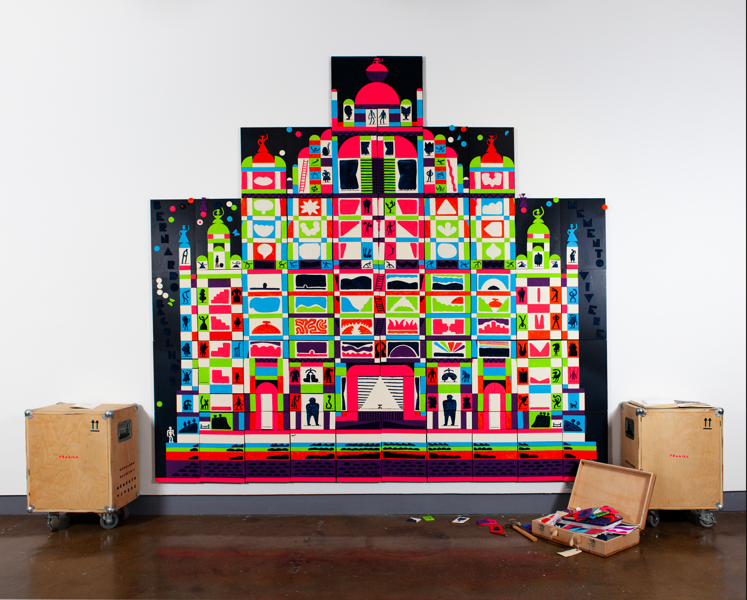 Colorful geometric artwork resembling a building on a white wall, with wooden crates and art supplies on the floor.