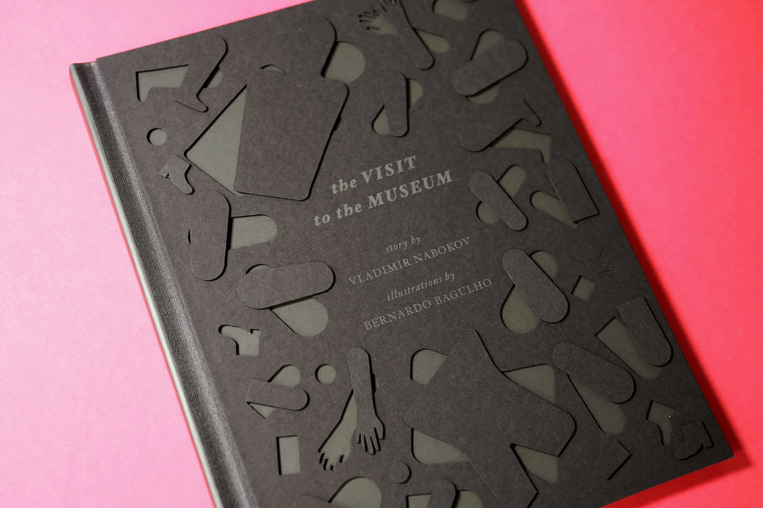 A black book with cut-out shapes of various objects and body parts on its cover, with the title 'the VISIT to the MUSEUM' and author's name Vladimir Nabokov, lie on a pink background.