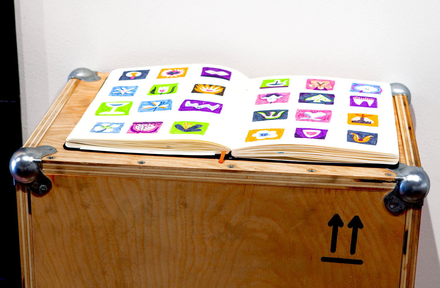 Open sketchbook with colorful painted squares on a wooden table with metal corner protectors, against a white wall.