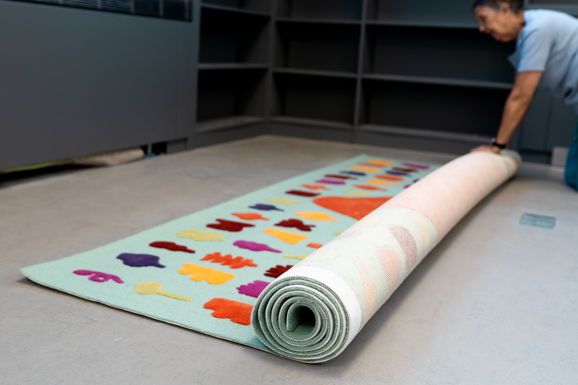 A person rolling out a colorful felt fabric with animal shapes on it onto a gray floor, with empty black shelves in the background.