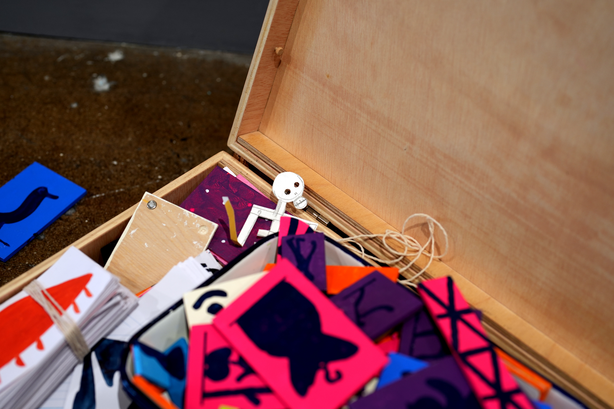 Open wooden box filled with colorful paper cards and paper cutouts with writing, along with some string and a talking dog figurine. A blue card with a black object is on the dark speckled surface nearby.
