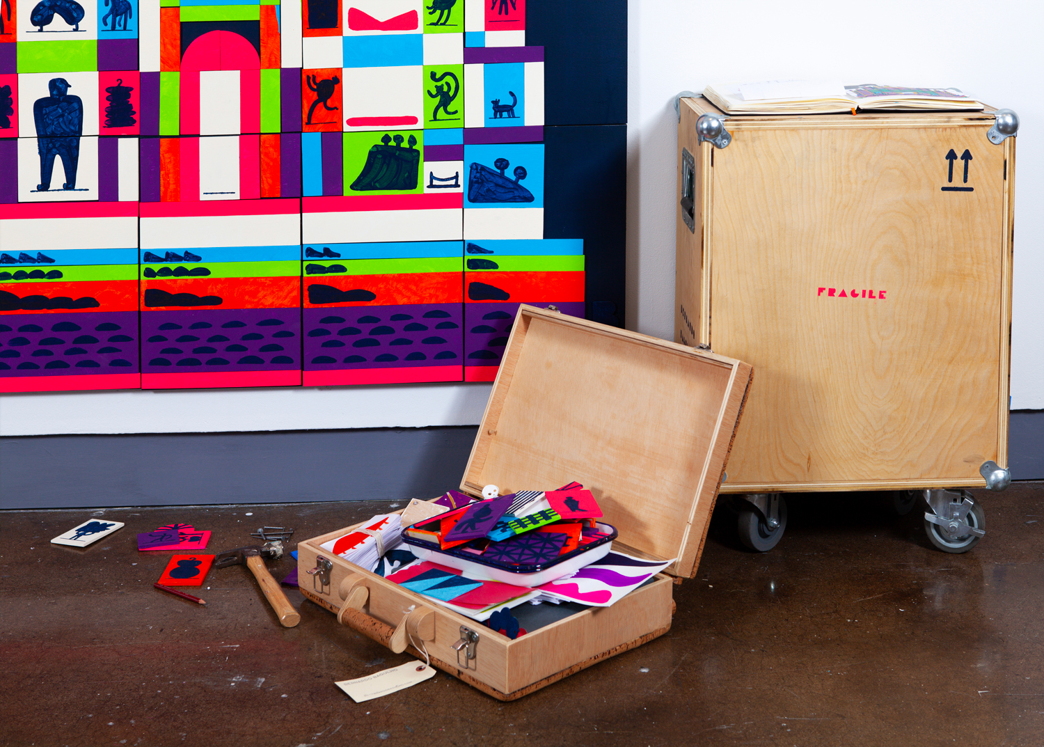 A colorful artwork on a wall made of stacked boxes with silhouettes of various figures. In front are open wooden crates and scattered art supplies, including papers, cutting tools, and colorful printed materials, on a brown floor.