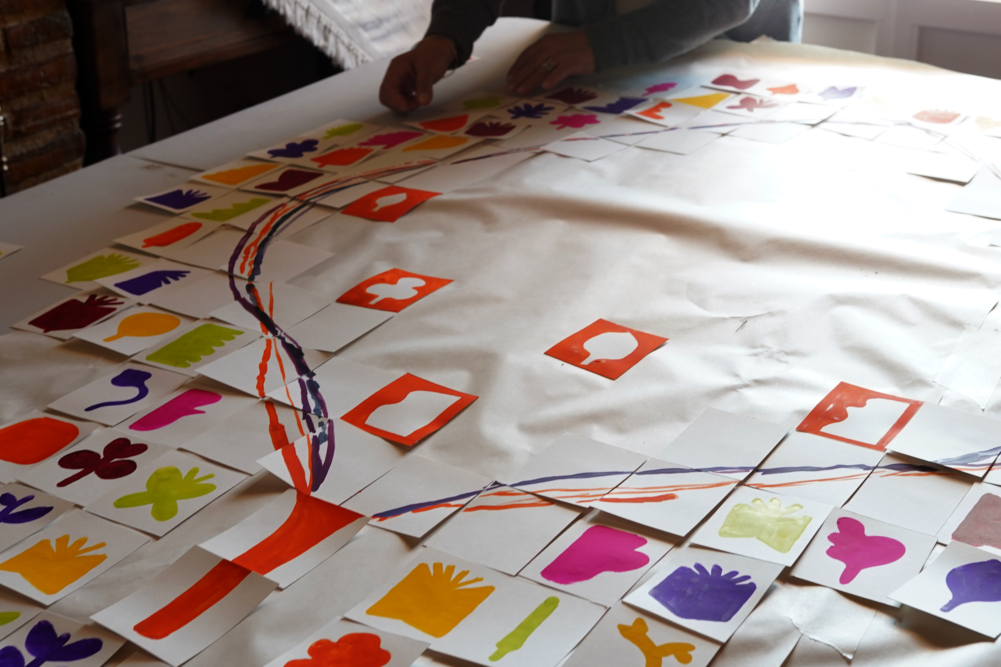 A person arranging colorful paper cutouts of various shapes, including hearts, flower-like shapes, and other abstract forms, on a large sheet of paper or fabric, with additional markers or paint drawn on the surface.