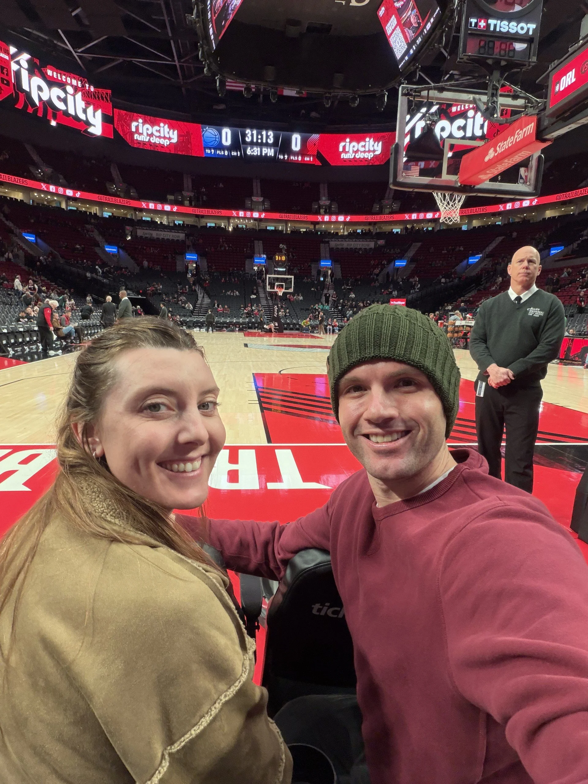 Two smiling people taking a selfie inside a basketball arena with an empty court, scoreboard, and seating in the background.