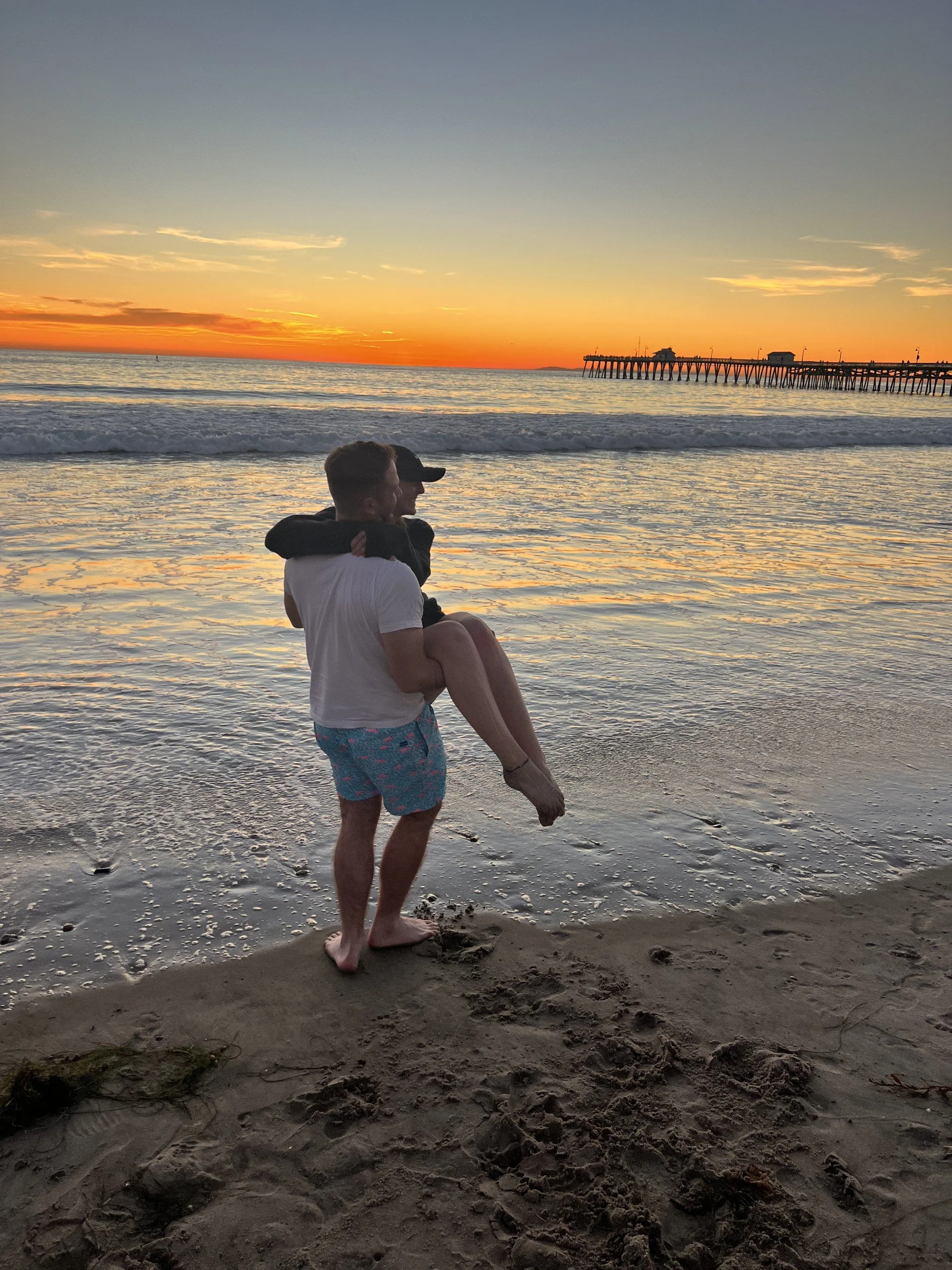A man carrying a woman on the beach at sunset, with a pier in the distance.