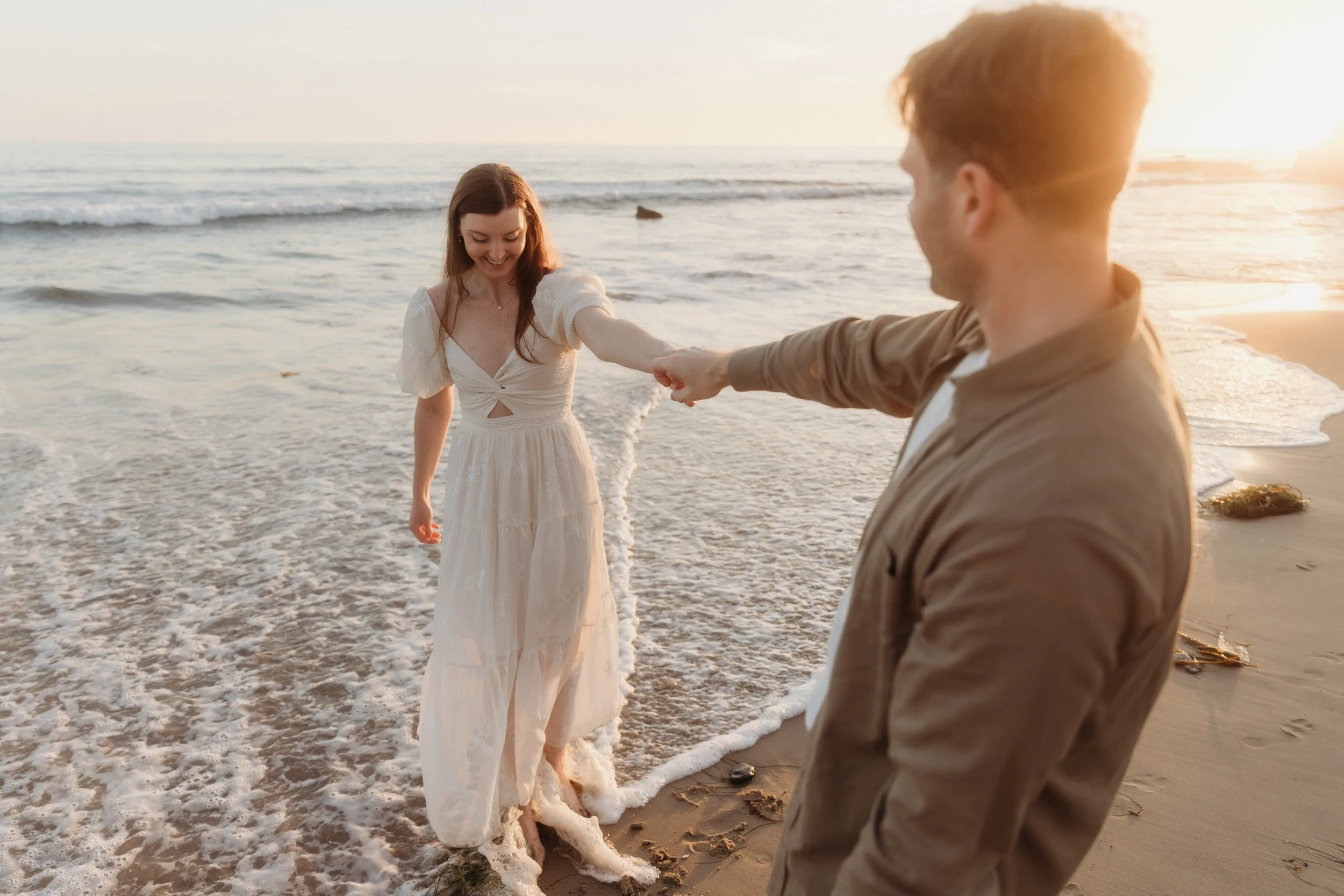 A couple on the beach during sunset, with the man touching the woman's hand as she steps into the water, both smiling.