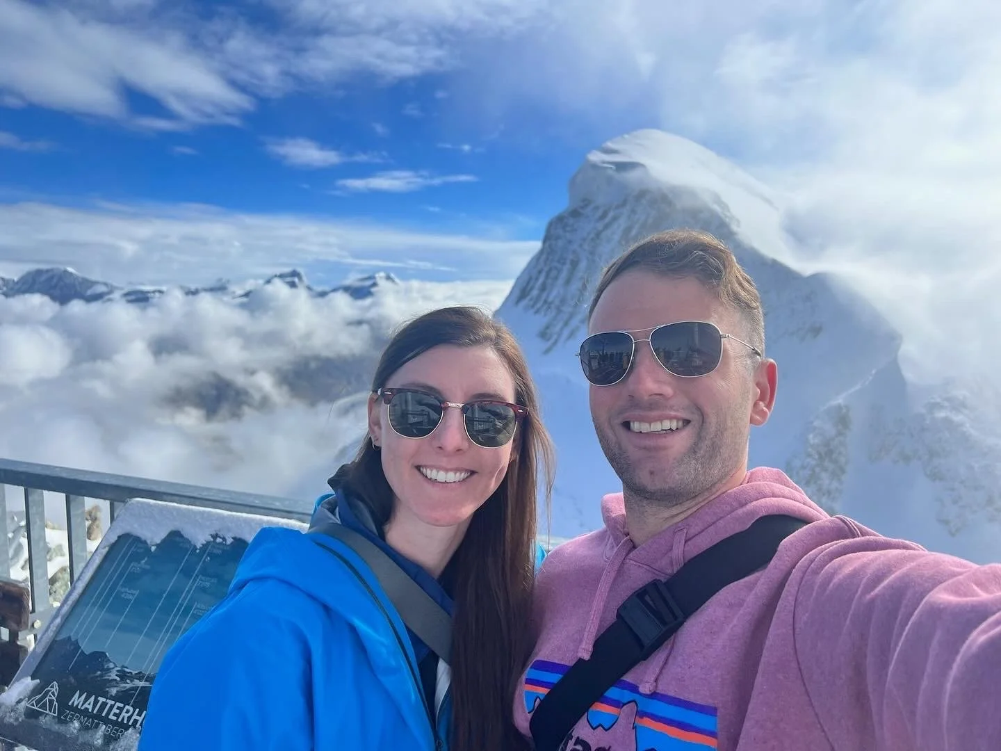 A smiling couple taking a selfie on a snowy mountain peak with snow-covered mountains in the background under a partly cloudy sky.
