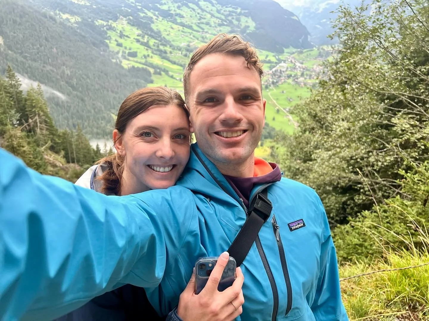 A smiling couple taking a selfie outdoors with a scenic mountain landscape in the background.