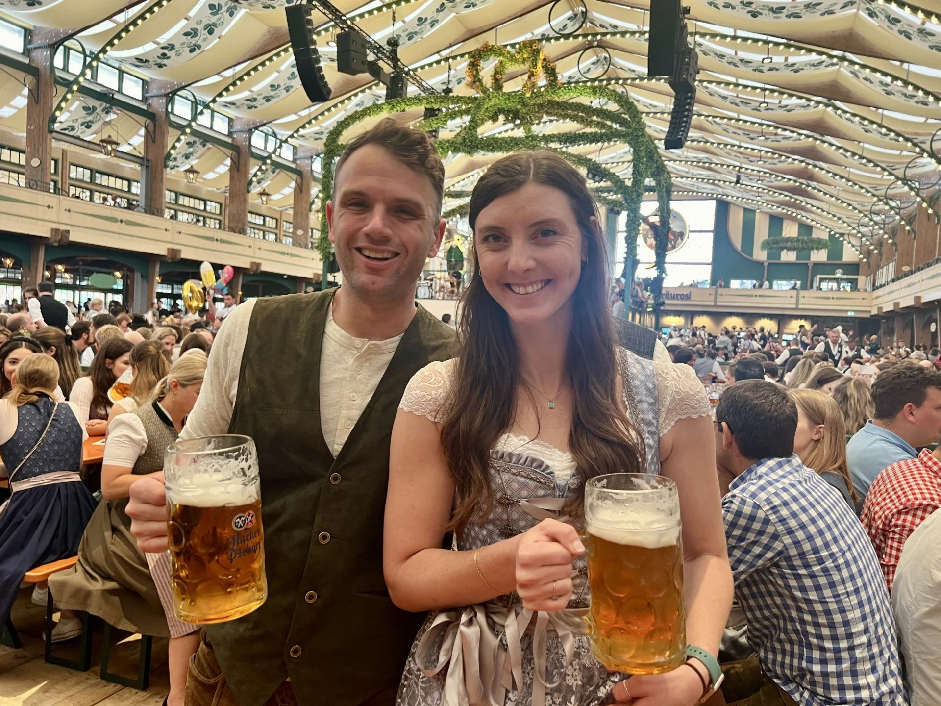 Two people smiling and holding mugs of beer at a large indoor festival or event with many people seated at long tables. The venue has high ceilings with lights and decorative greenery.