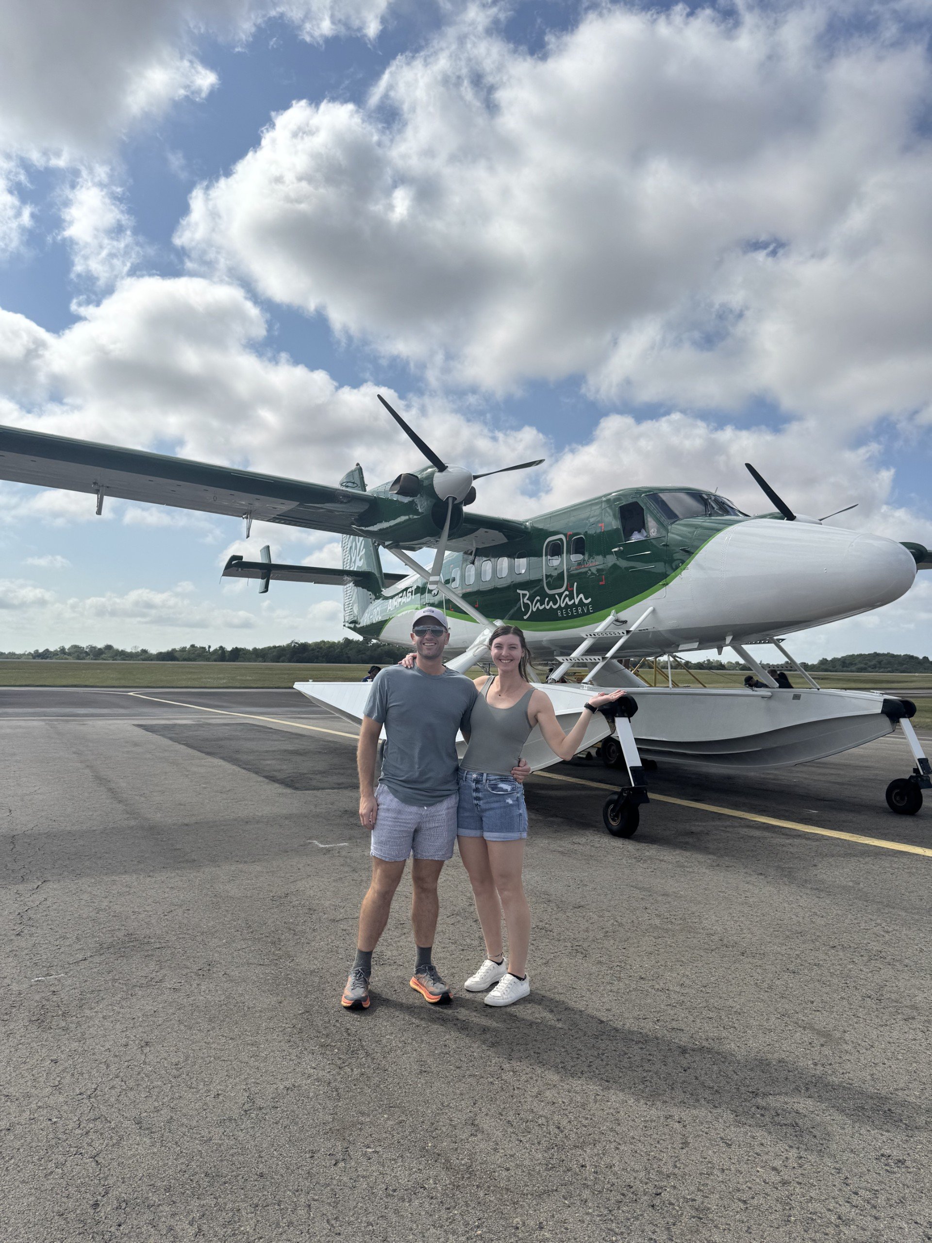 A man and woman standing together in front of a seaplane on a runway, smiling at the camera, with clouds in the sky.