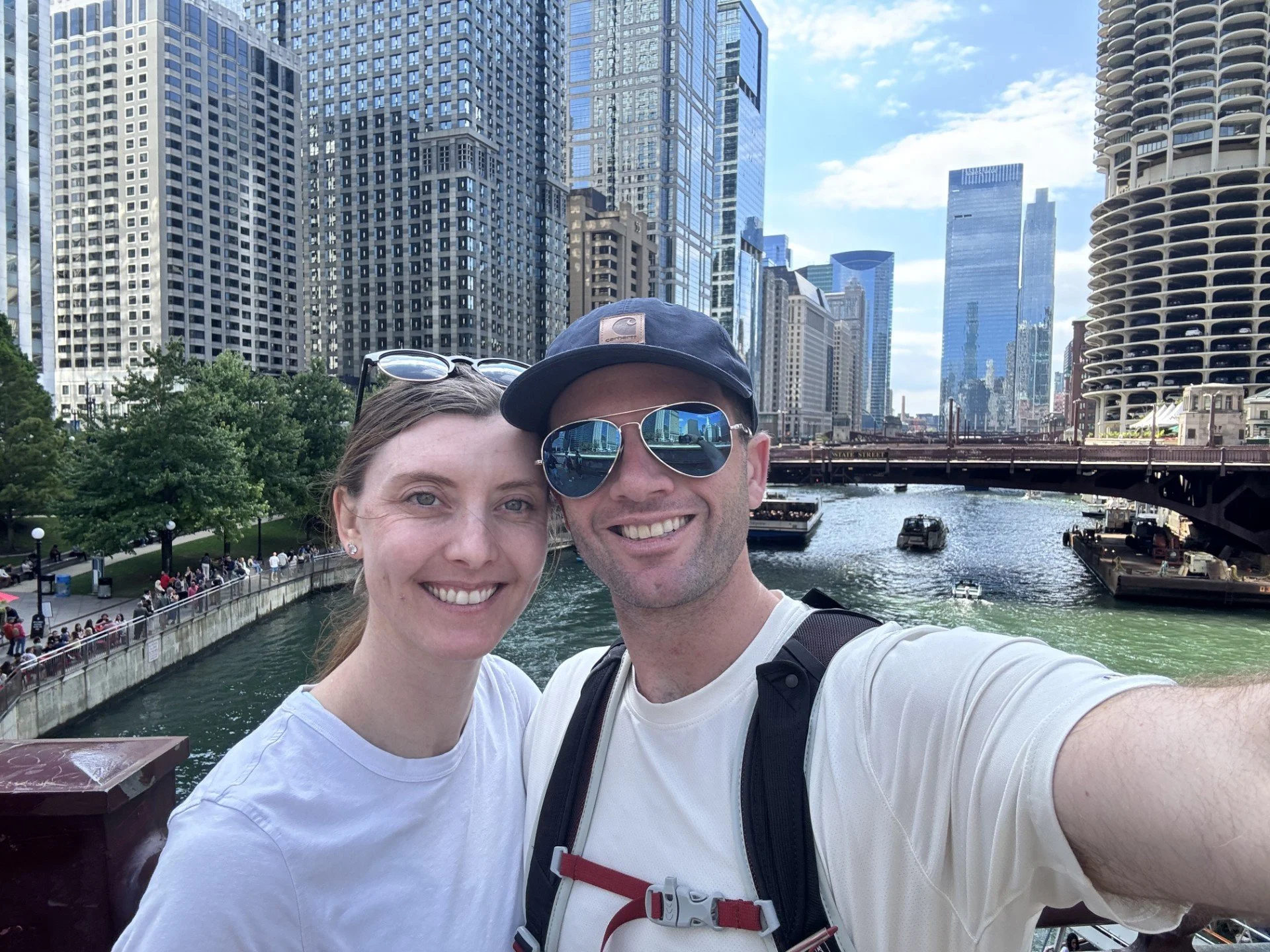 Smiling couple taking a selfie in front of a cityscape with tall buildings and a river.