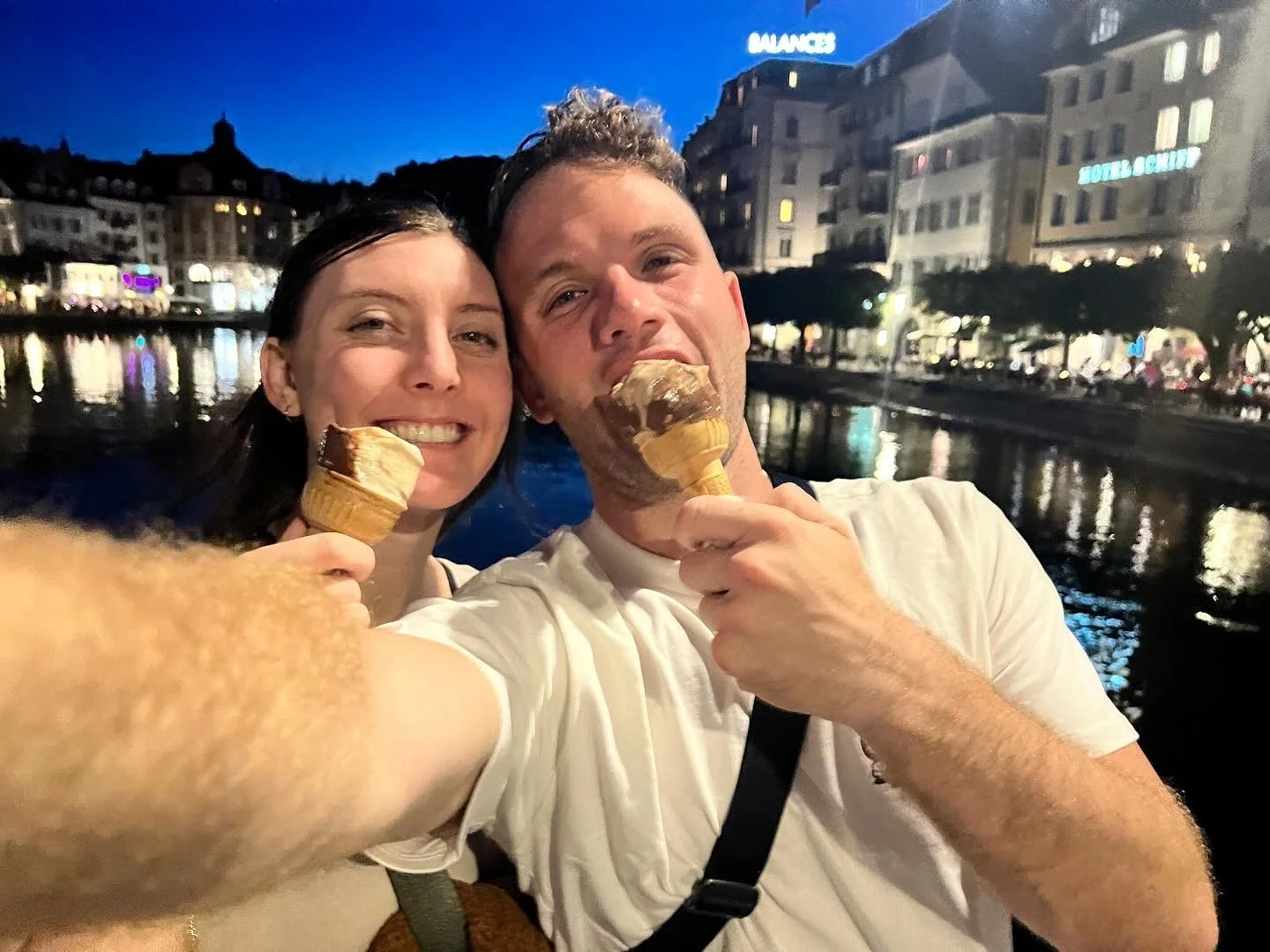 A couple taking a selfie while enjoying ice cream cones near a waterfront at night, with buildings lit up in the background.