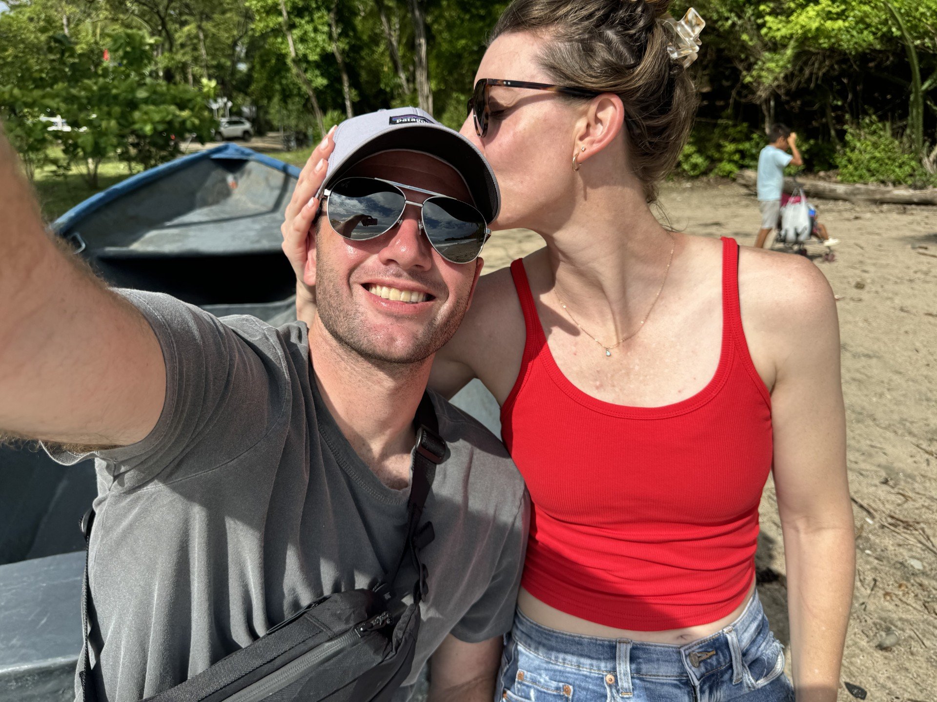 A man and woman taking a selfie on a beach, with a woman in the background near trees and a stroller.