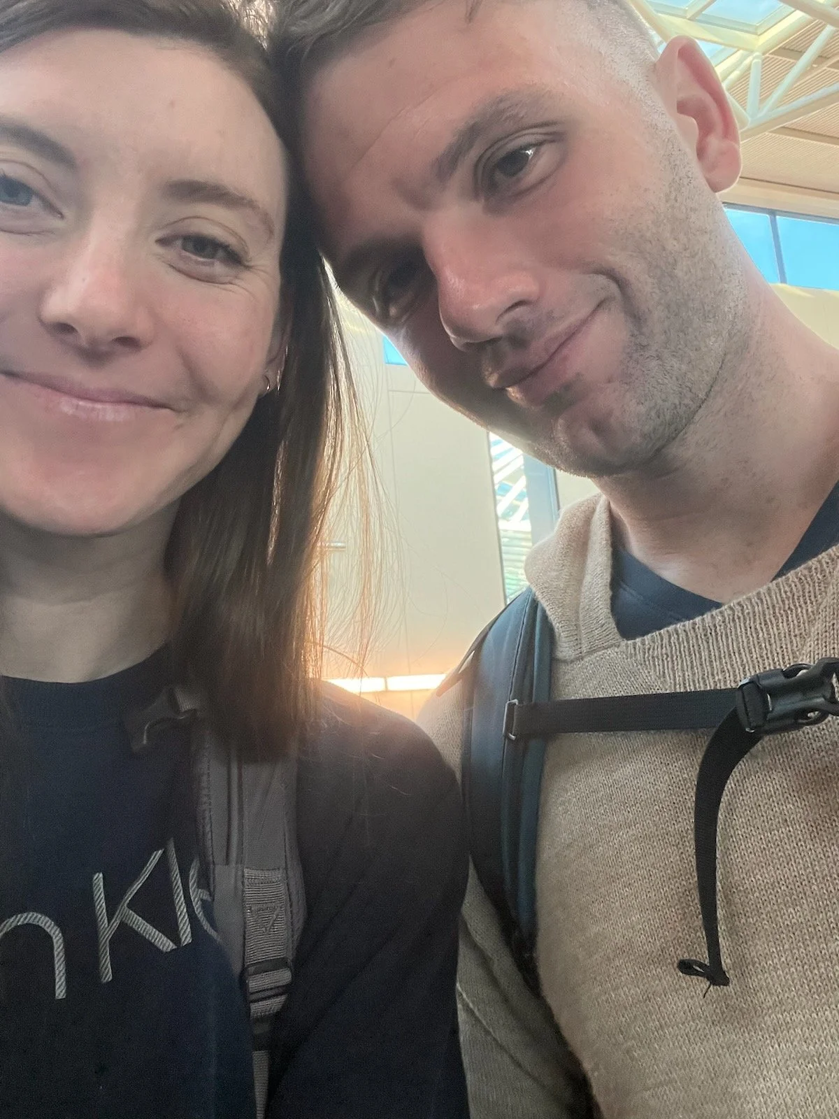 A smiling woman and a man taking a close-up selfie at an indoor location, with blue windows and metal roof structures visible in the background.