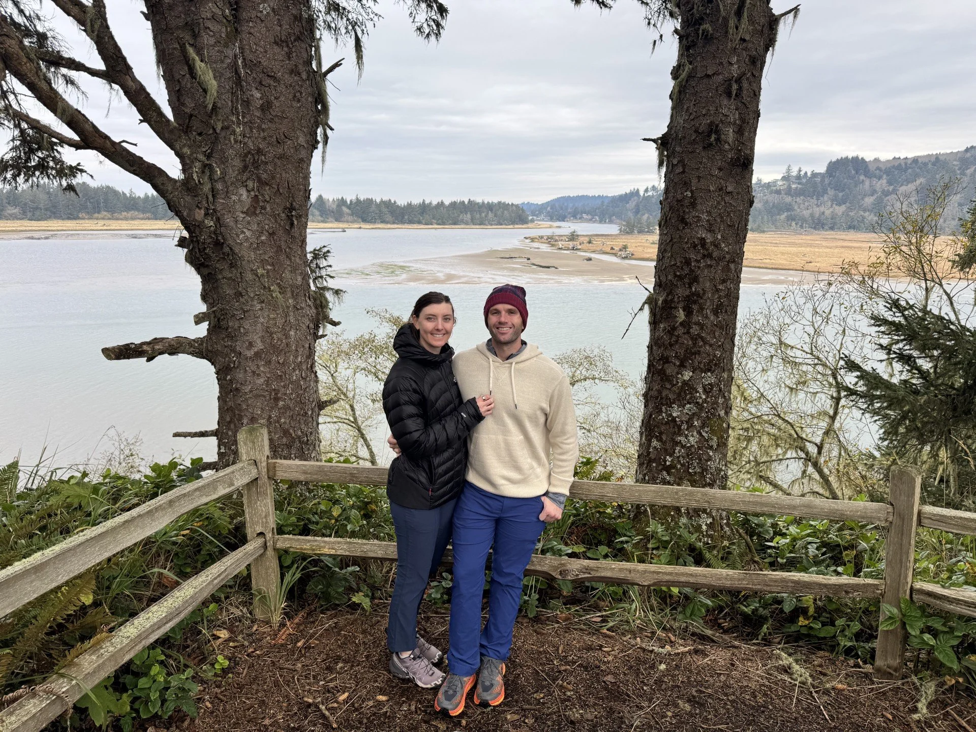 A smiling couple standing close together in outdoor clothing, surrounded by trees and overlooking a body of water with a cloudy sky.