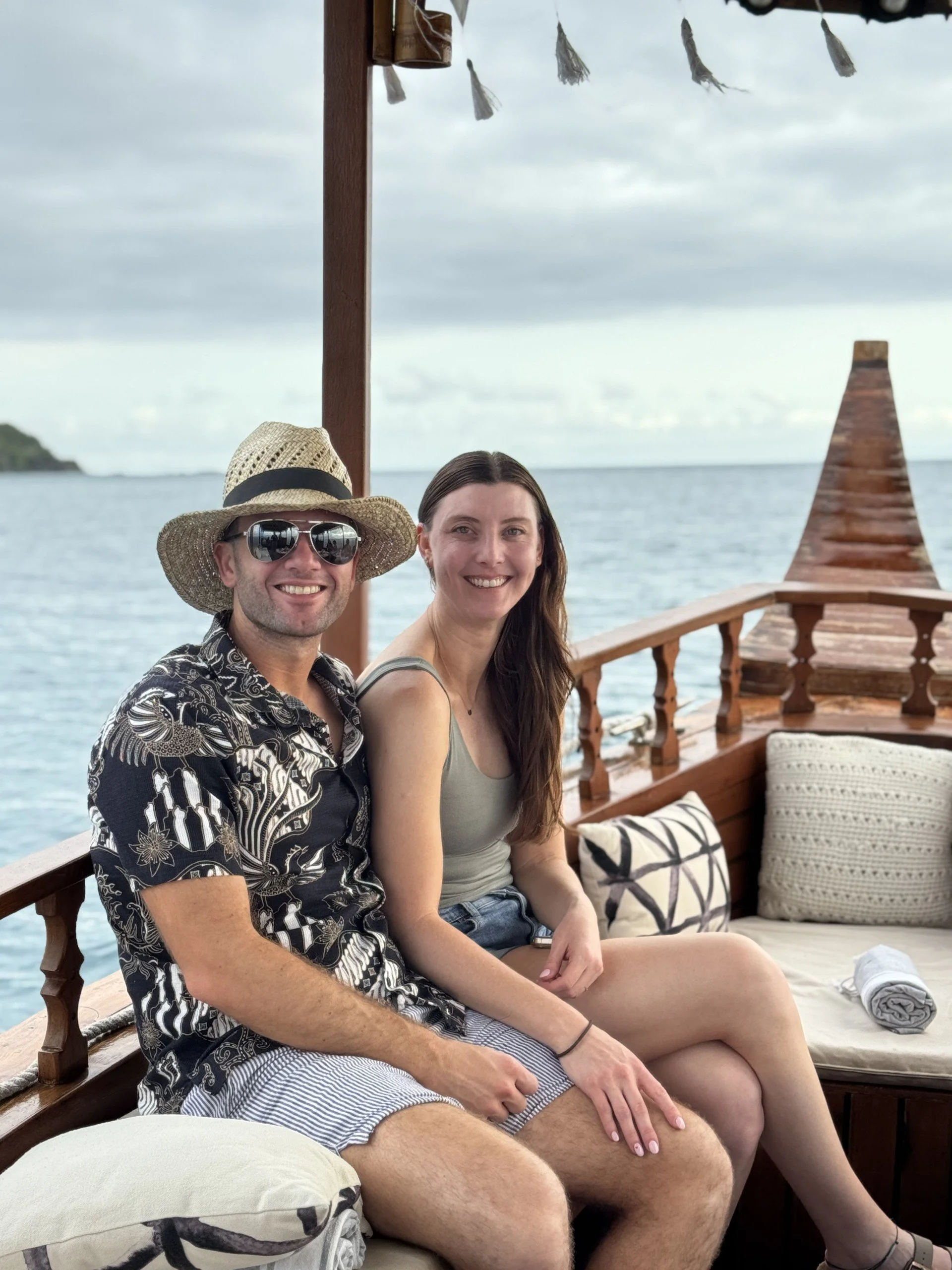 A smiling man wearing sunglasses, a straw hat, and a tropical shirt, sitting next to a smiling woman with long brown hair, on a boat with a wooden railing, cushions, and an ocean view in the background.