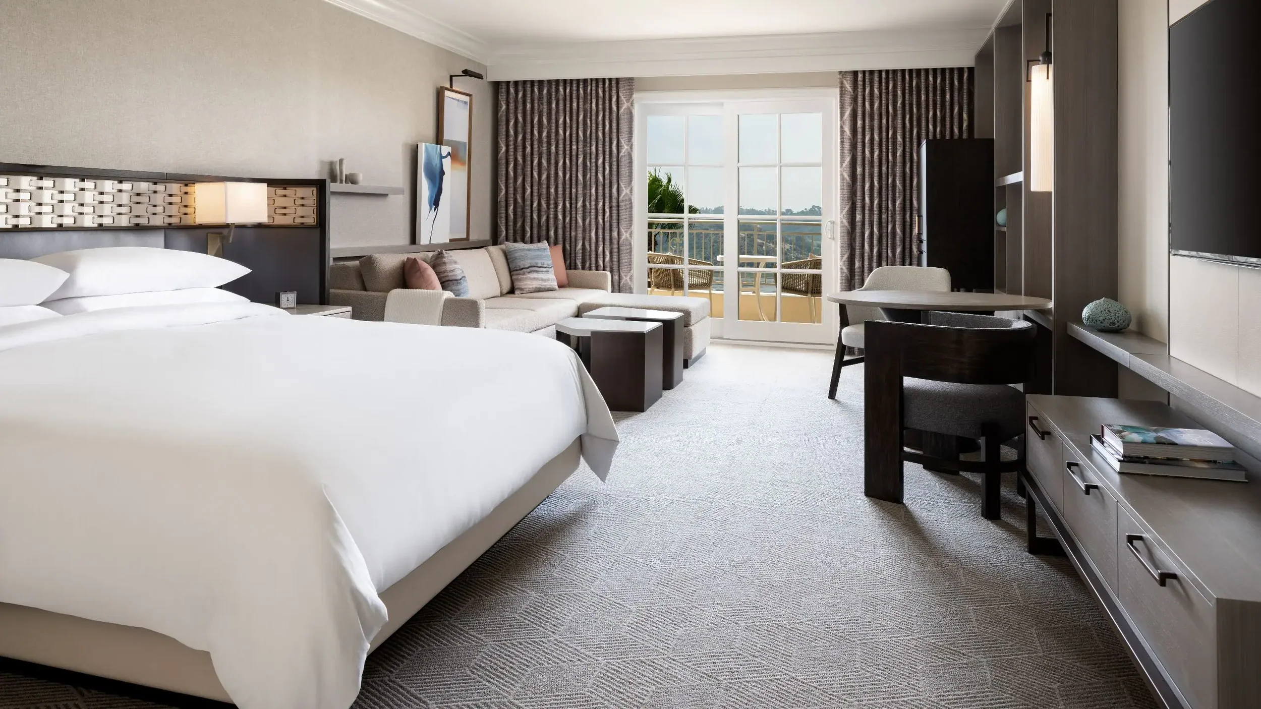 A hotel room with a large bed, a sitting area with a sofa, a balcony with outdoor seating, a flat-screen TV, and modern furniture and decor.