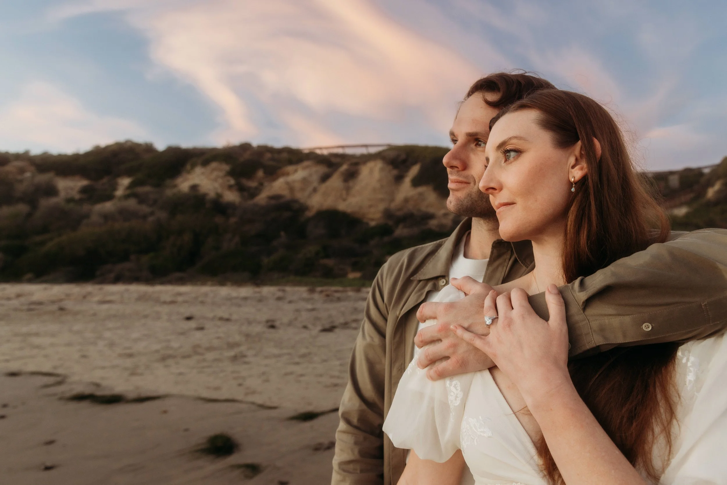A couple stands on a beach during sunset, with the man embracing the woman from behind. They are looking into the distance, with the woman's hand on her chest showing an engagement ring. The background features cliffs and a colorful sky.
