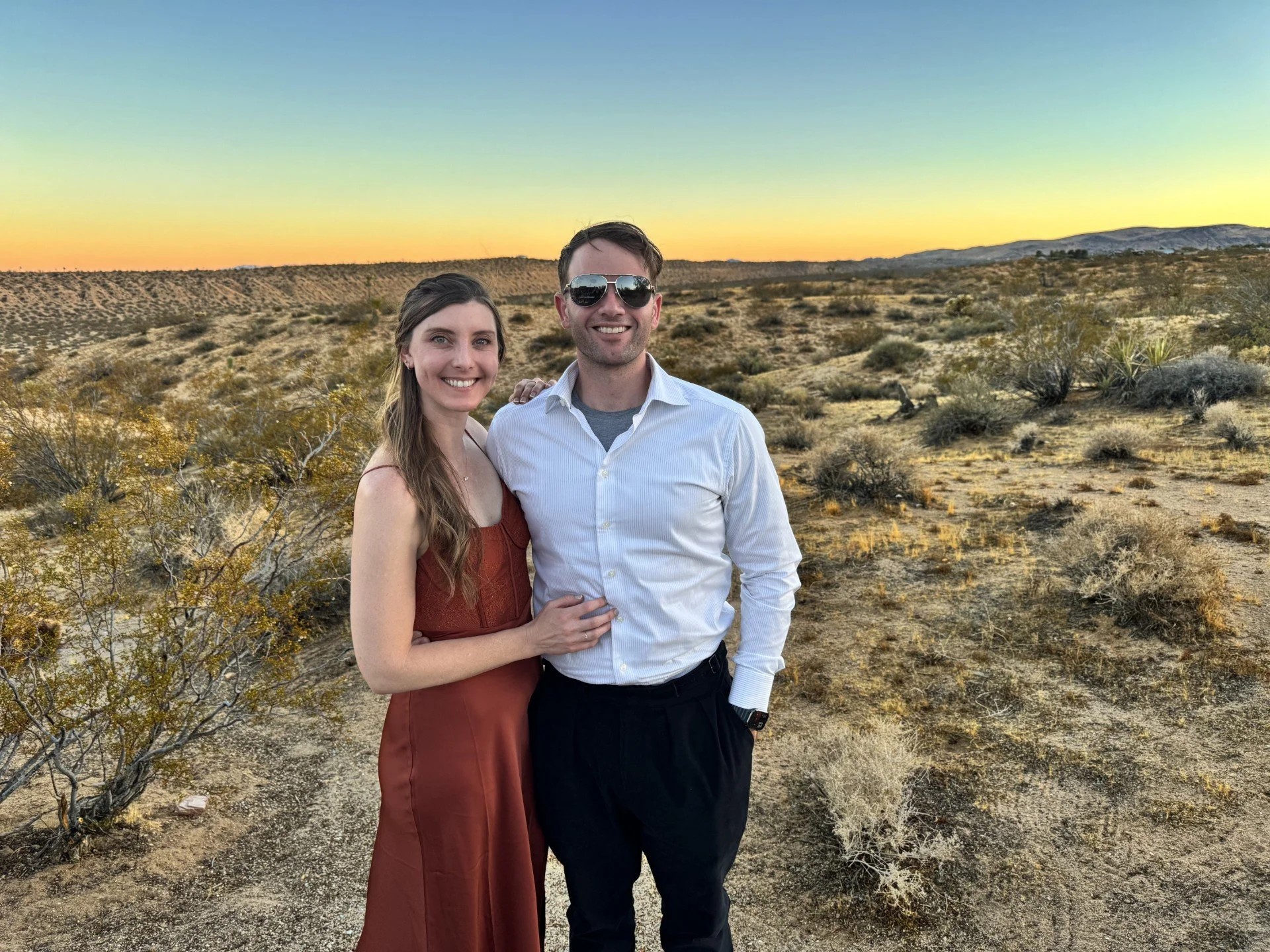 A smiling couple stands together in a desert landscape during sunset, with sparse bushes and distant hills under a colorful sky.