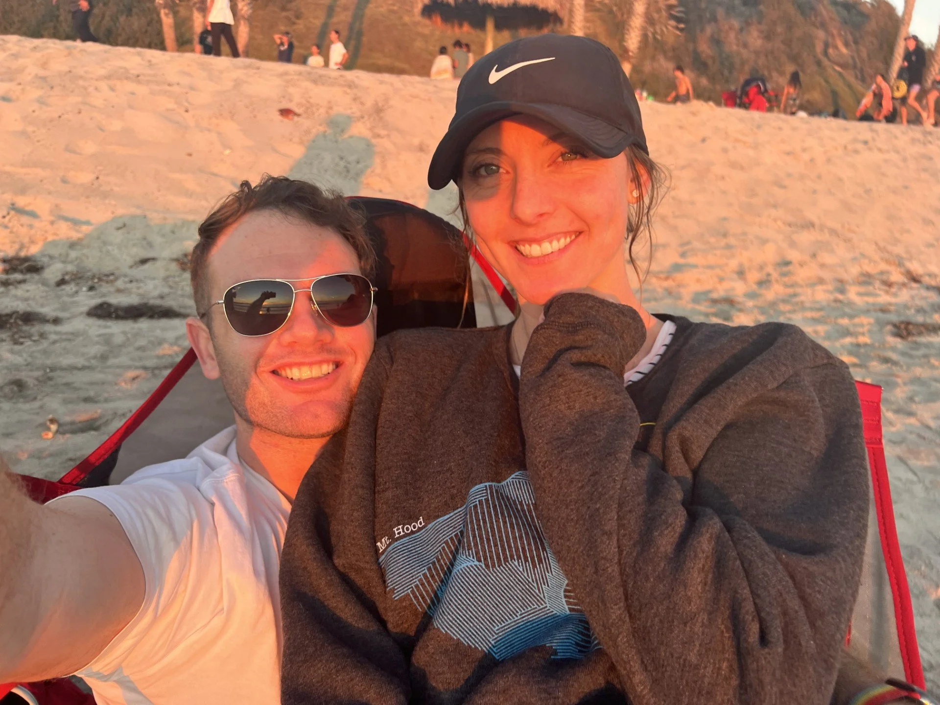 A smiling couple taking a beach selfie during sunset with sandy dunes and other beachgoers in the background.