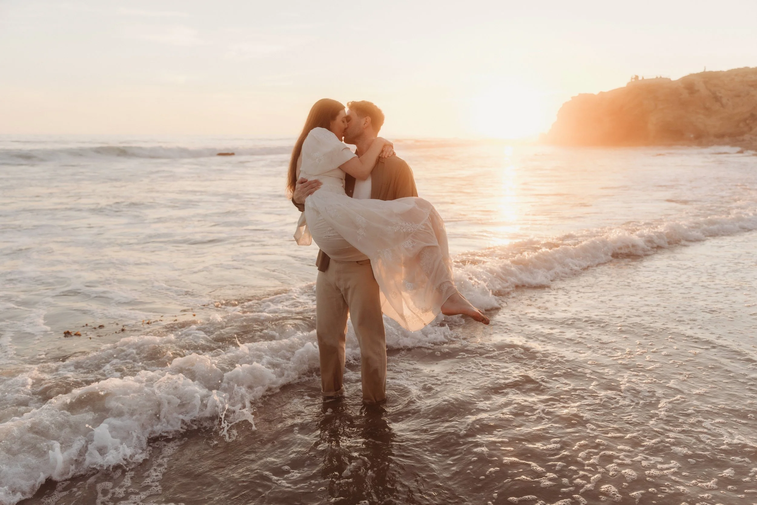 A couple on the beach during sunset, with the man holding the woman in his arms, both sharing a kiss, ocean waves at their feet, and a rocky cliff in the background.