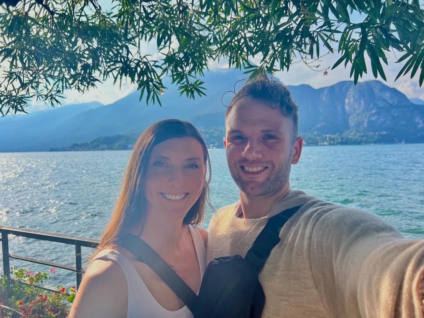 A smiling couple taking a selfie by a lake with mountains in the background, partially shaded by overhanging branches.