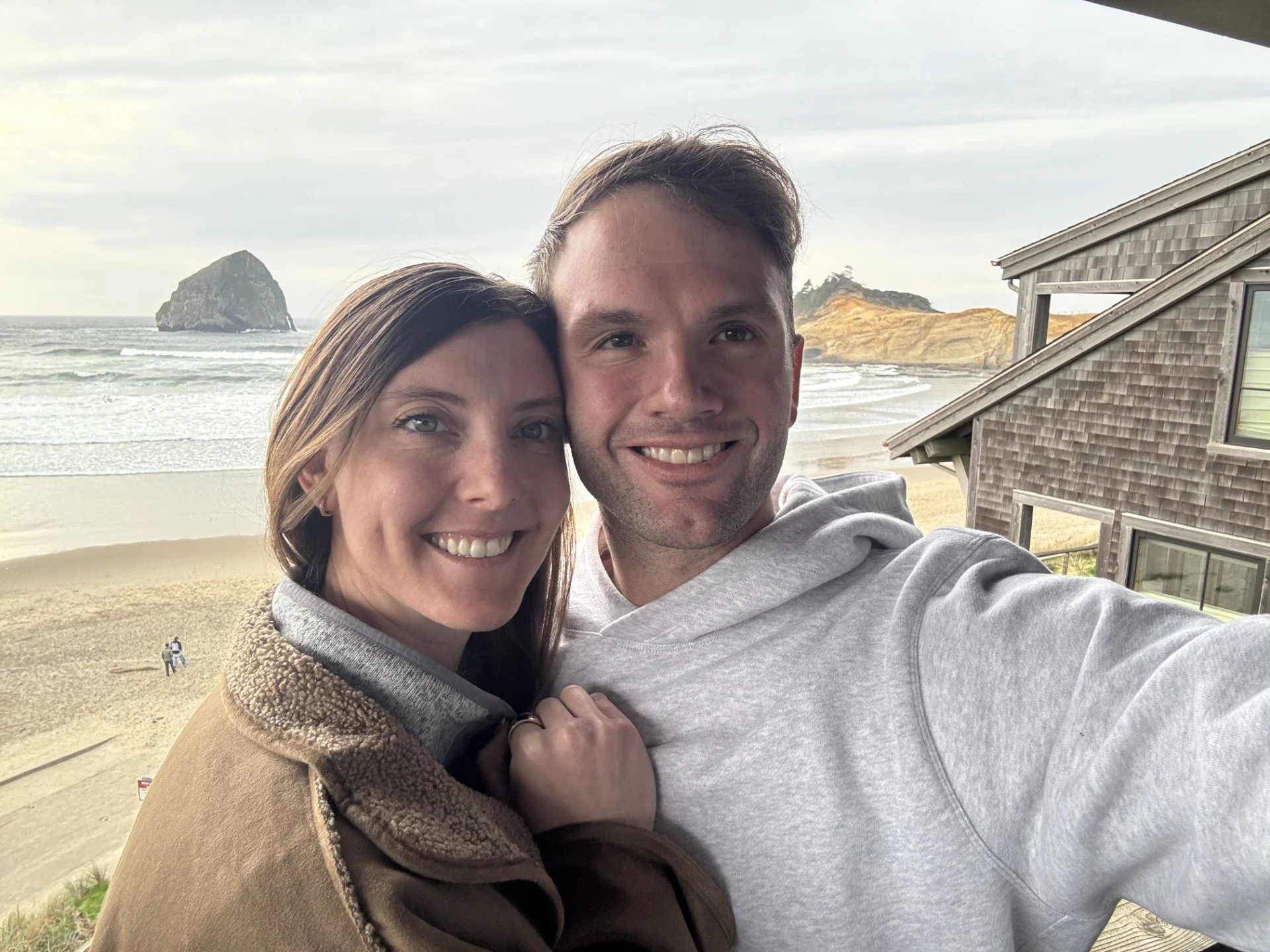 A smiling couple taking a selfie on a beach with ocean waves, a large rock formation, and hills in the background.