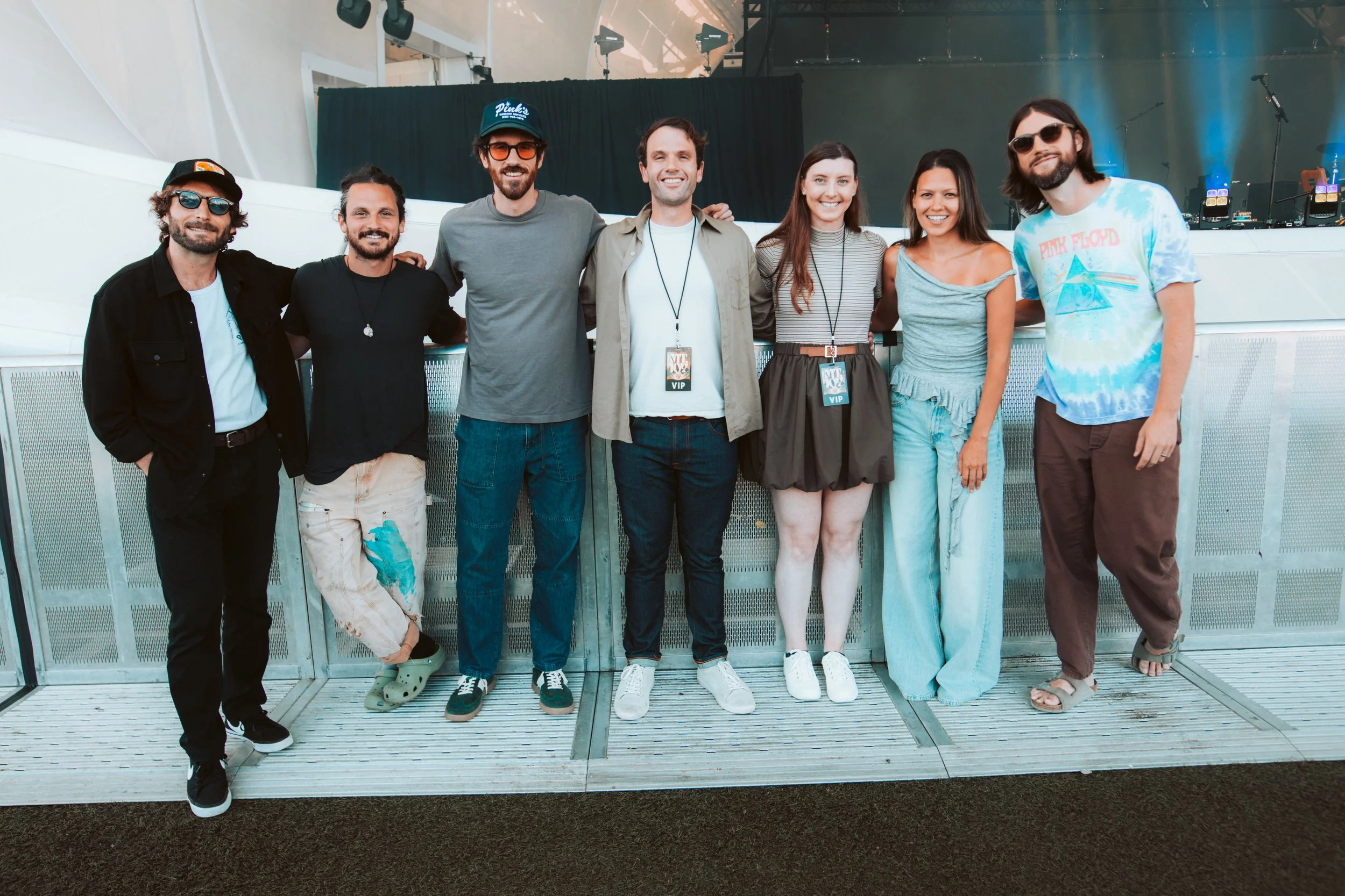 Group of seven friends standing together at an indoor event, smiling for the camera, with a stage in the background.