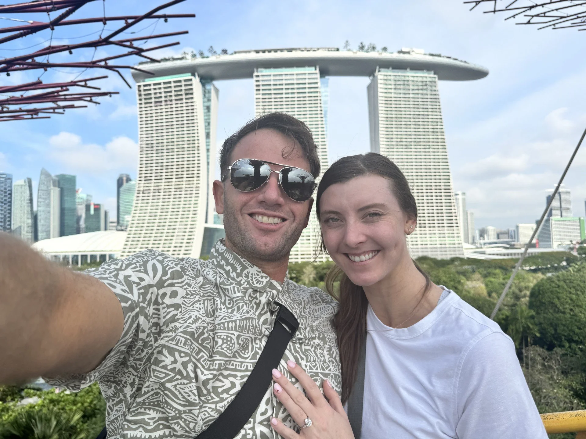 A smiling couple taking a selfie in front of the Marina Bay Sands hotel in Singapore. The man is wearing sunglasses and a patterned shirt, and the woman is smiling with her hand on the man's chest, showing an engagement ring.
