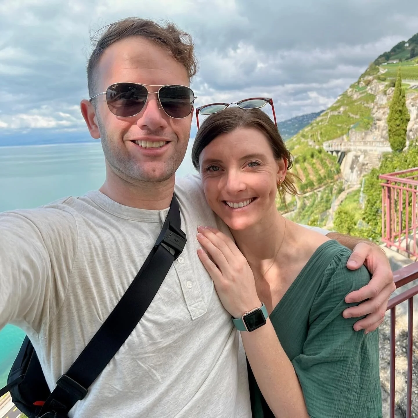 A smiling couple taking a selfie at a scenic outdoor location with a body of water and green hills in the background.