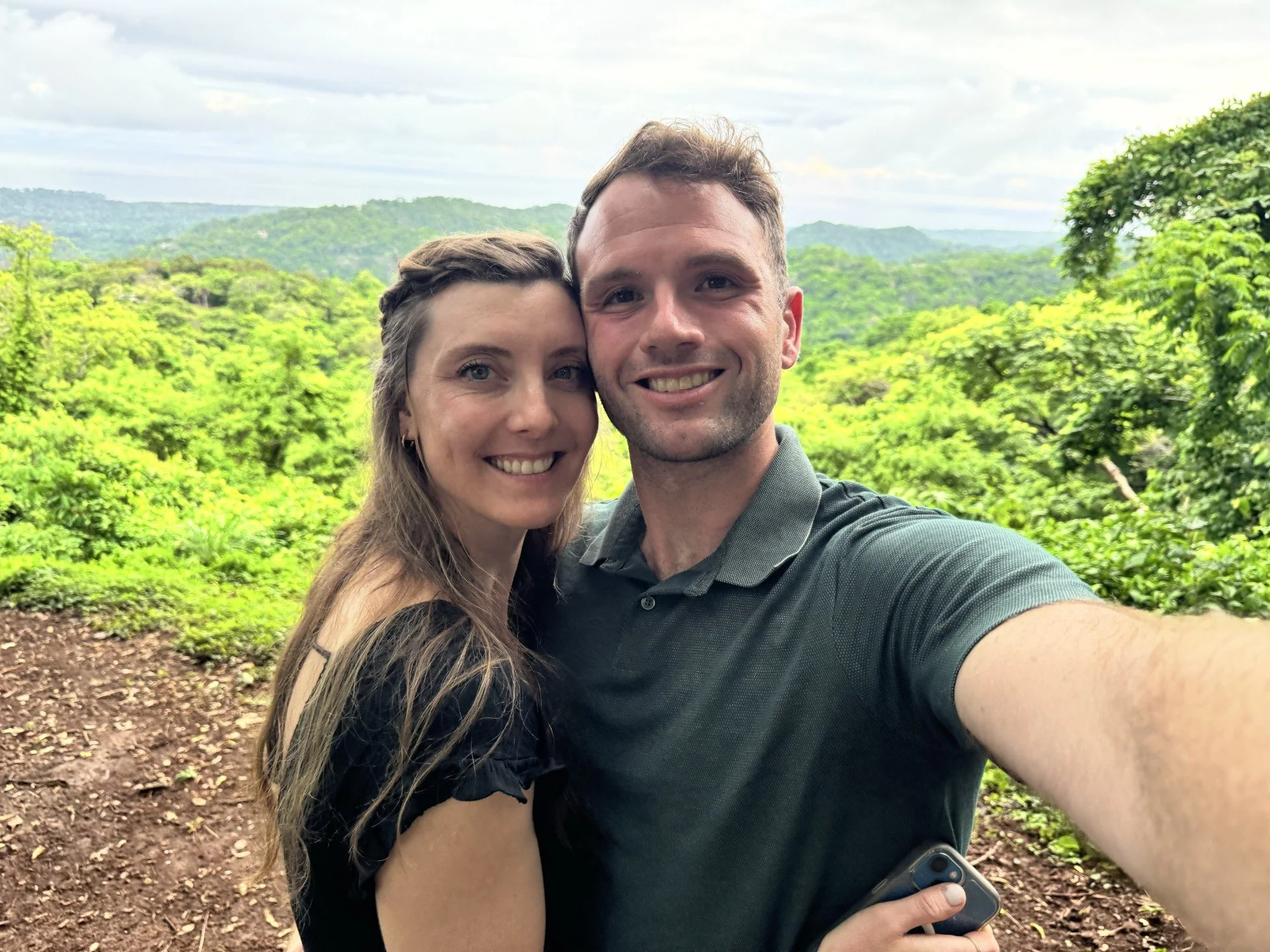 A smiling couple taking a selfie in front of lush green trees and rolling hills with cloudy sky in the background.