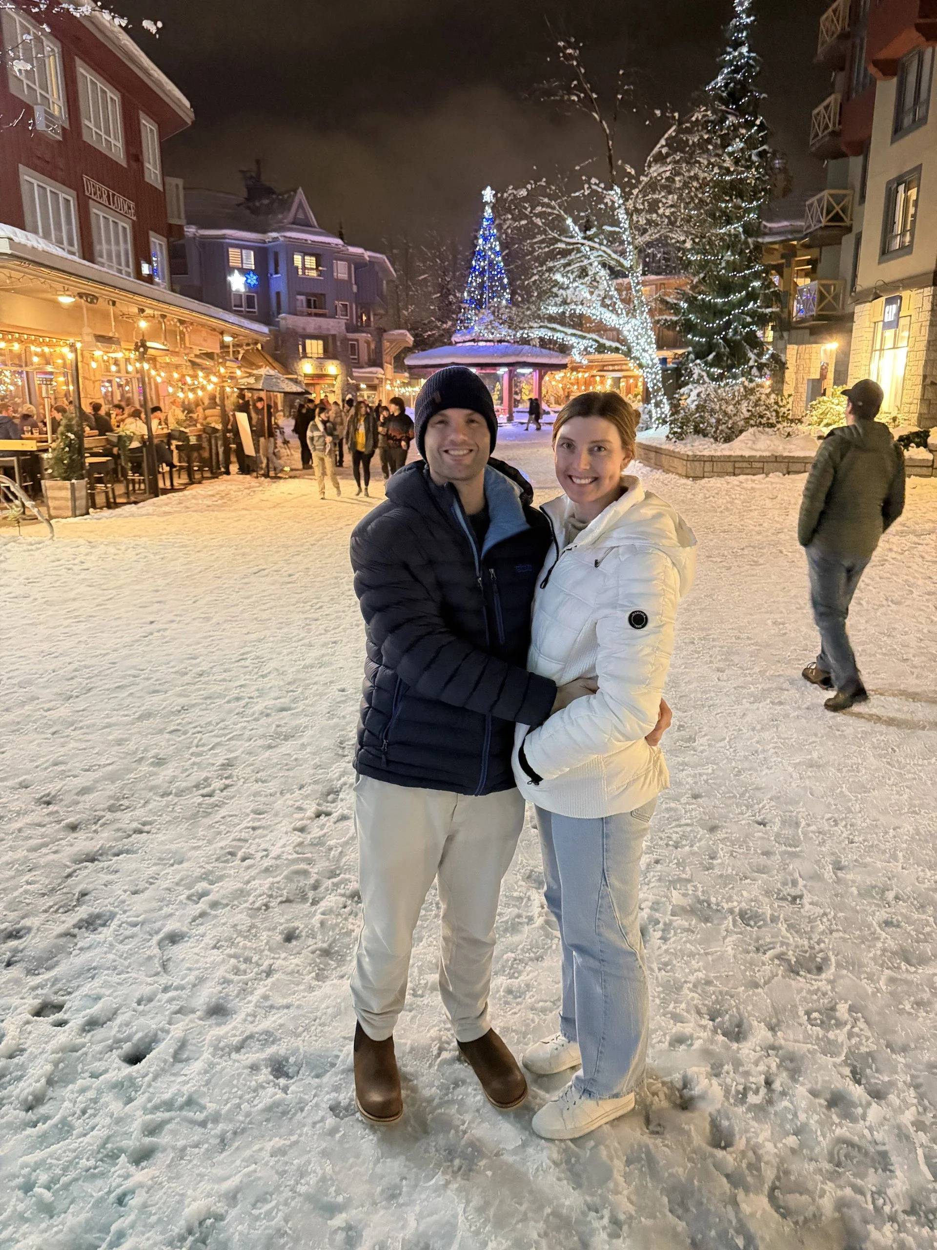 A smiling couple standing close together outdoors on a snowy night, with a festive holiday scene behind them including Christmas trees and string lights on buildings, and other people walking and dining in a lively winter setting.
