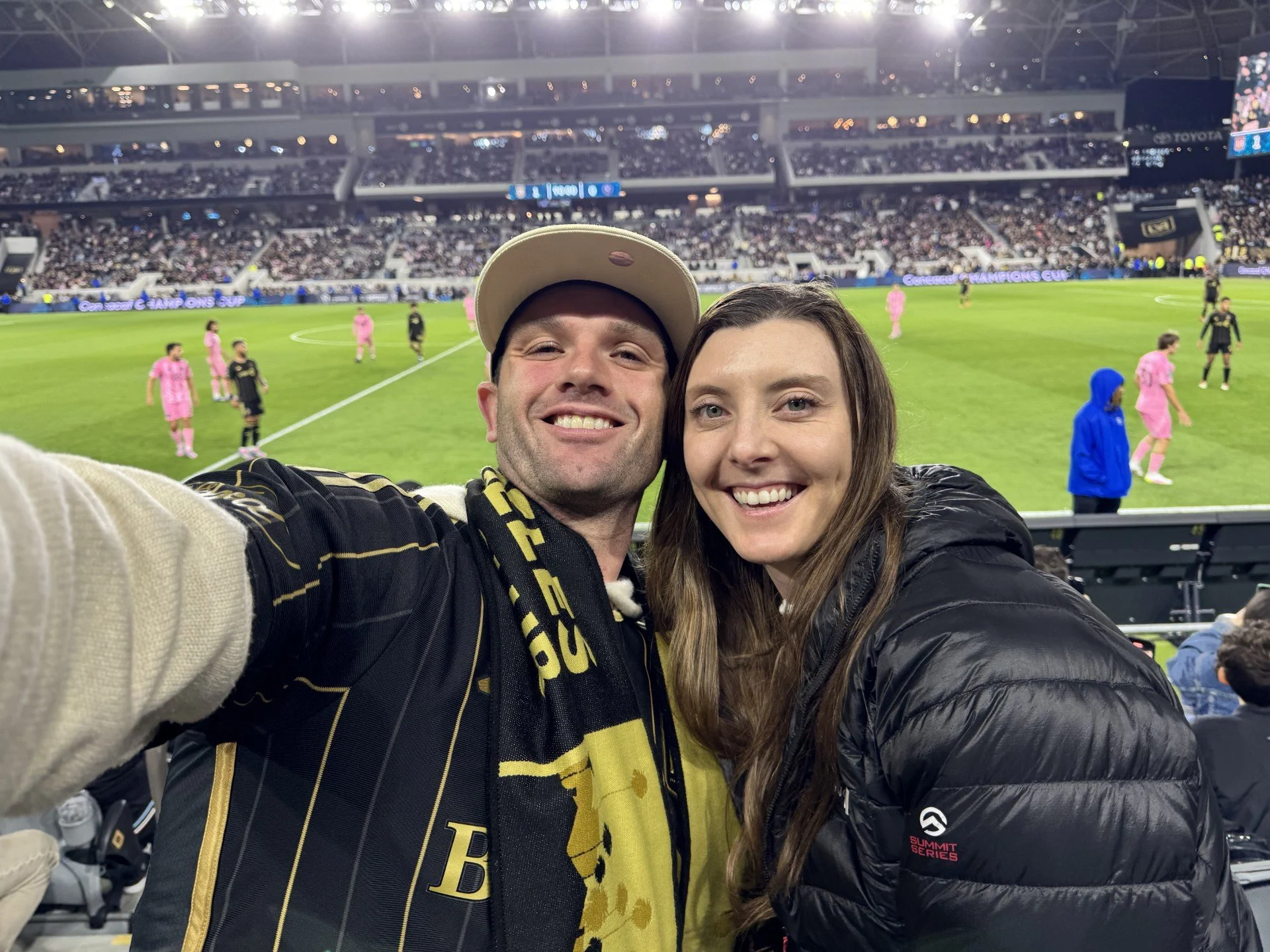 Two smiling people taking a selfie at a soccer stadium with players on the field in the background; the person on the left is wearing a beige cap and sports attire with a yellow and black scarf, and the person on the right has long brown hair and is 