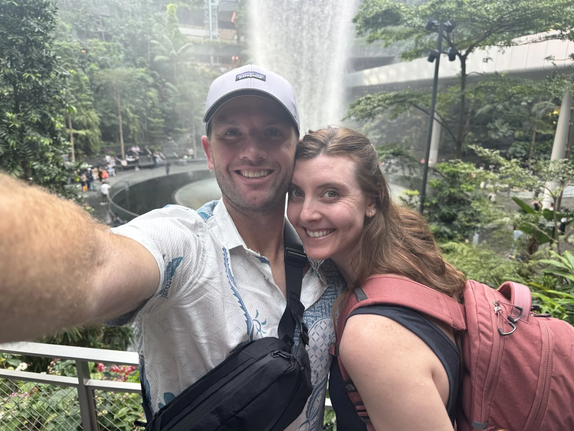A smiling couple taking a selfie outdoors near a waterfalls and lush greenery.