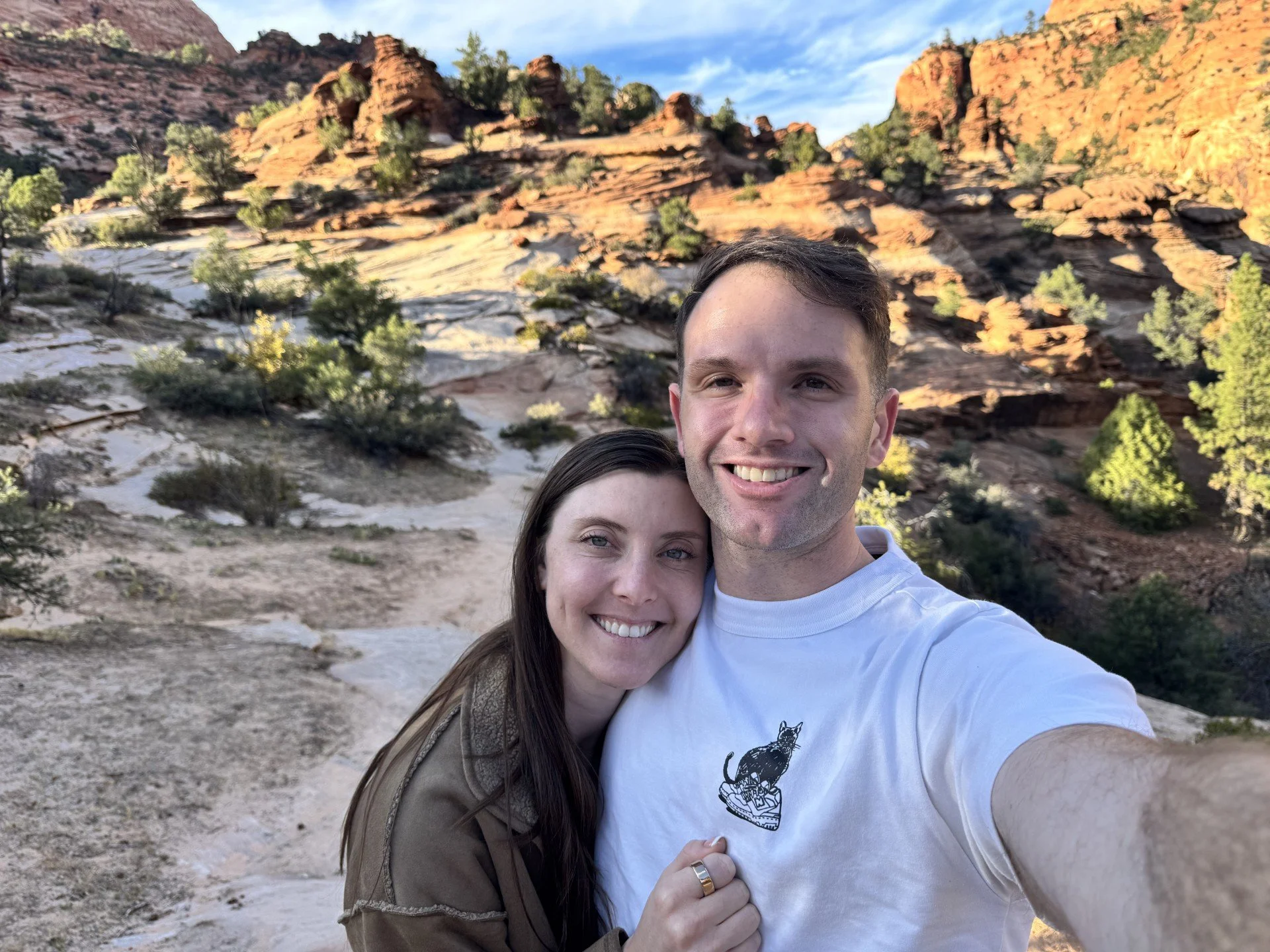 A smiling couple taking a selfie outdoors in a desert landscape with rocky hills and sparse green shrubbery in the background.