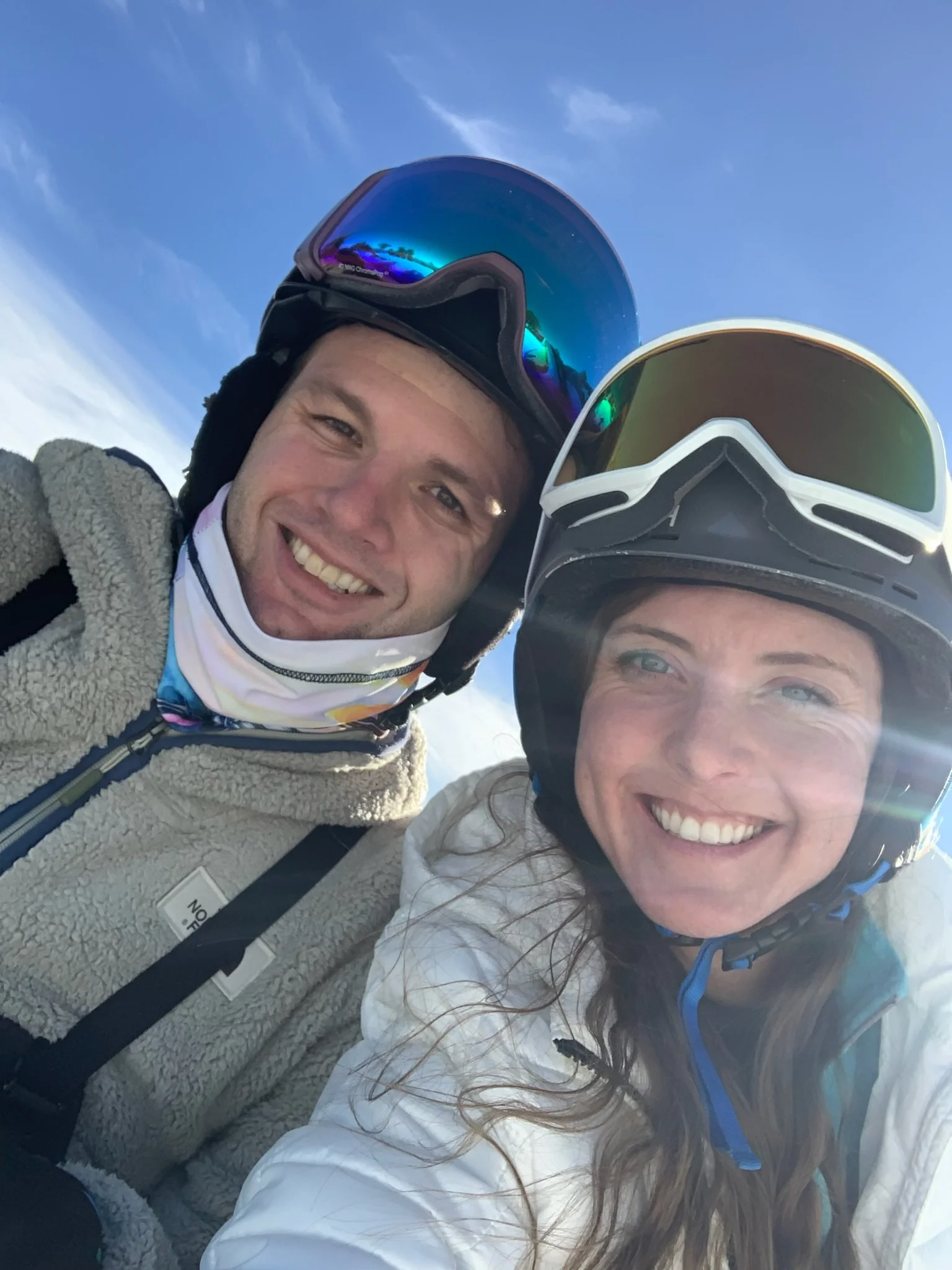 A man and a woman wearing ski helmets and goggles, smiling outdoors with a blue sky in the background.