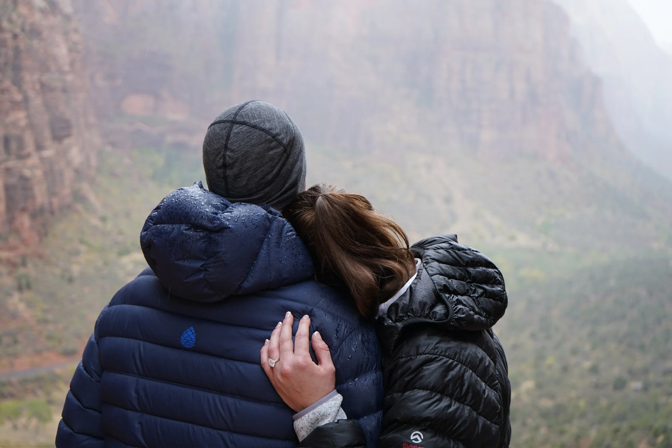 A couple embracing outdoors at a scenic canyon view on a foggy day, wearing warm jackets.