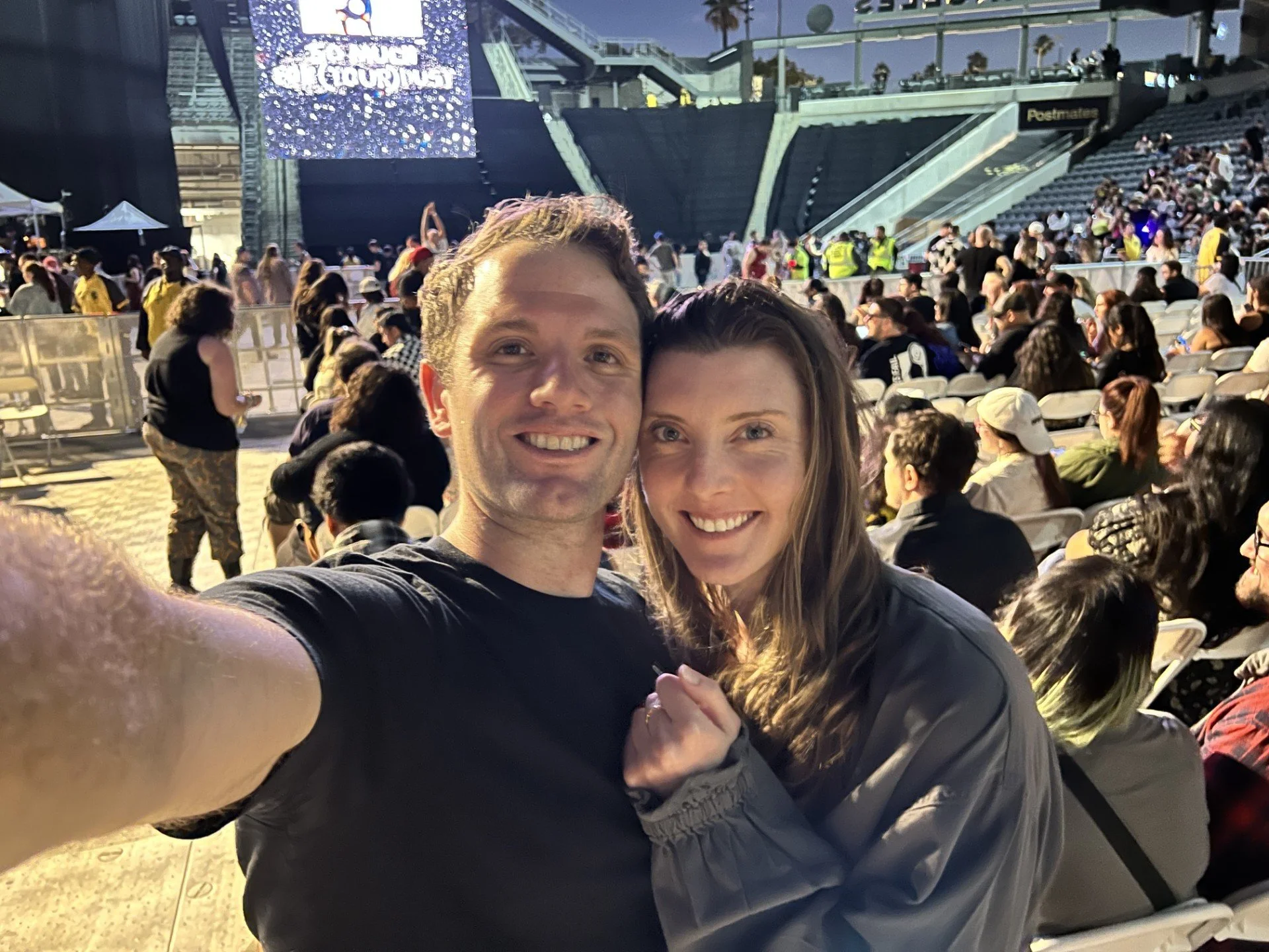 A smiling man and woman taking a selfie at an outdoor event or concert, with a large crowd seated behind them and a stage in the background.
